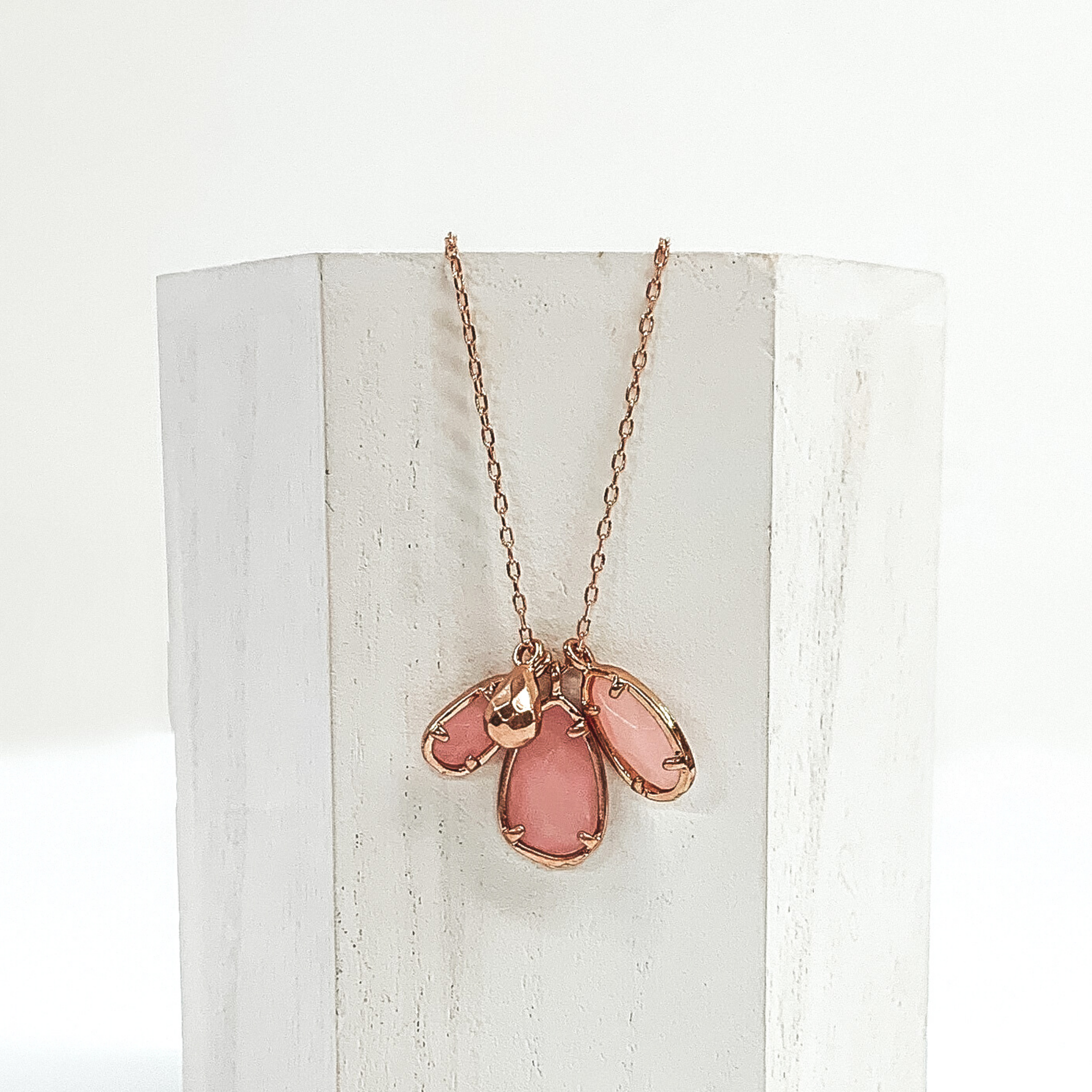 Small gold chain with three rose marbled teardrop stone pendants outlined in gold. It also has a tiny gold teardrop pendant. This necklace is pictured laying a white piece of wood on a white background.