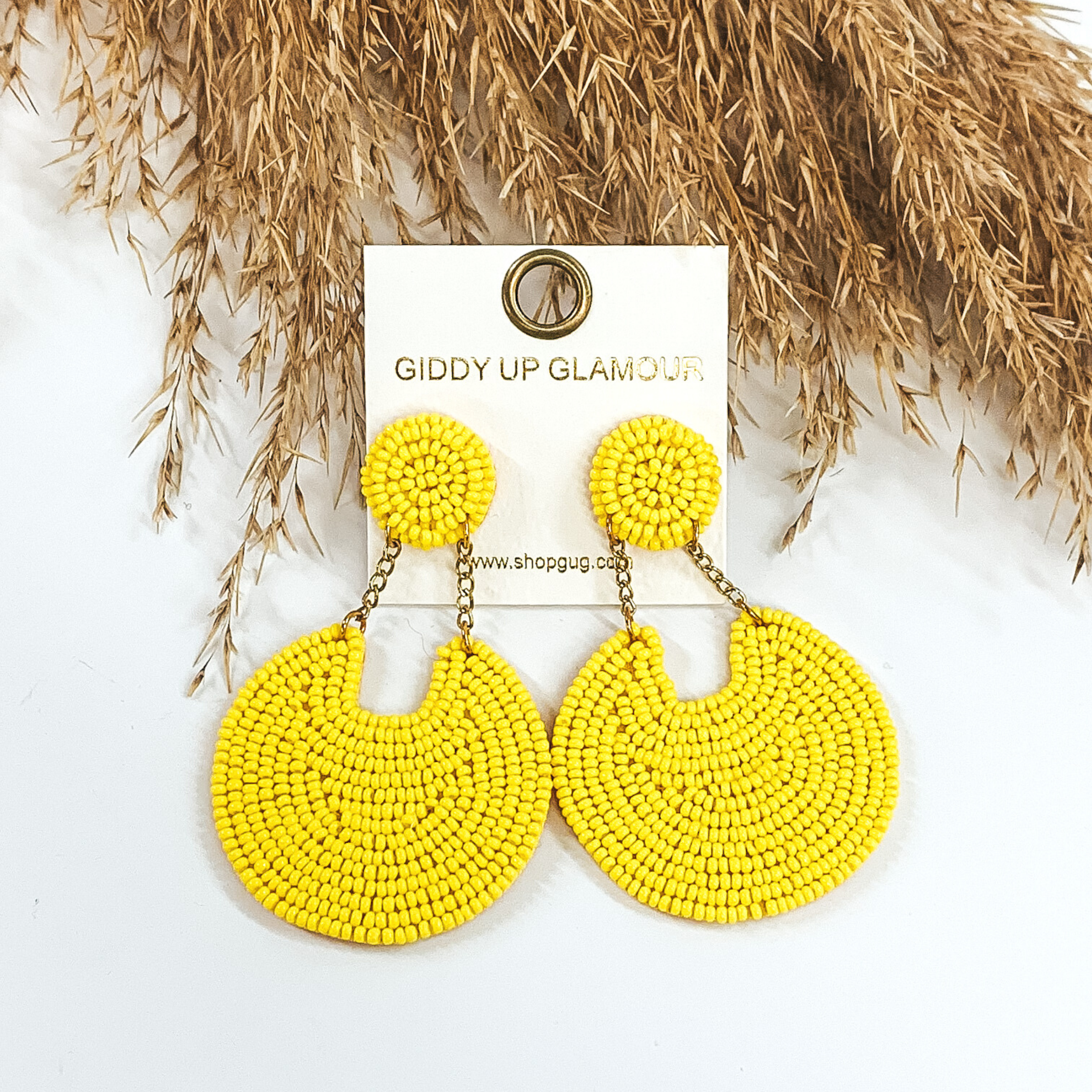 These earrings are covered in yellow beads. There is a circle stud with two gold chains that hold a bigger, hanging circle that has a notch out of the top. These earrings are pictured on a white background with tan floral at the top of the picture.