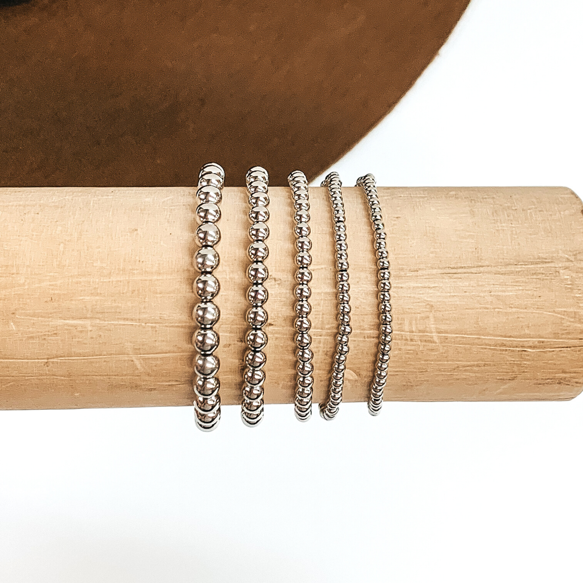 Set of five silver beaded bracelets in different sizes. These bracelets are pictured on a wooden bracelet holder on a white and brown background. 