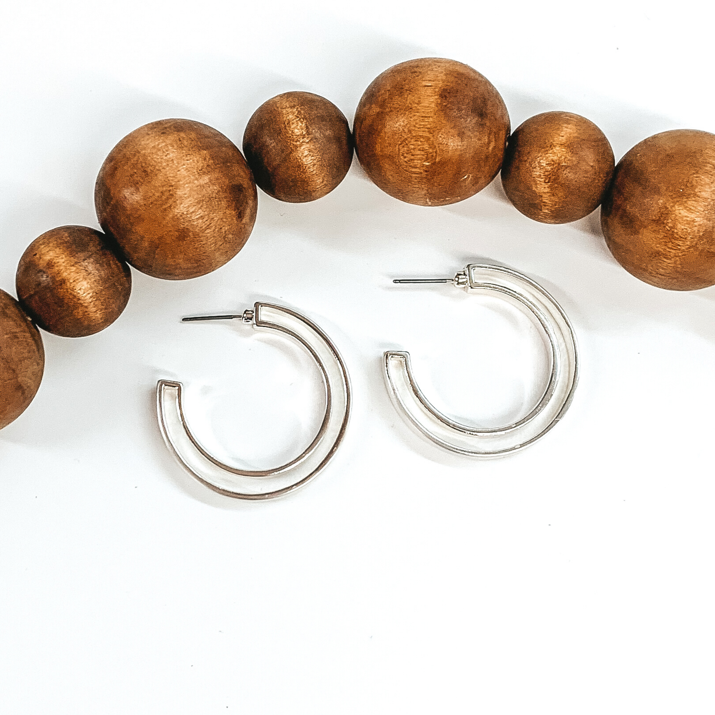 Laying on a white background are two silver hoop earrings. They are a circle double layered hoop earrings. There are brown decor beads above the earrings. 