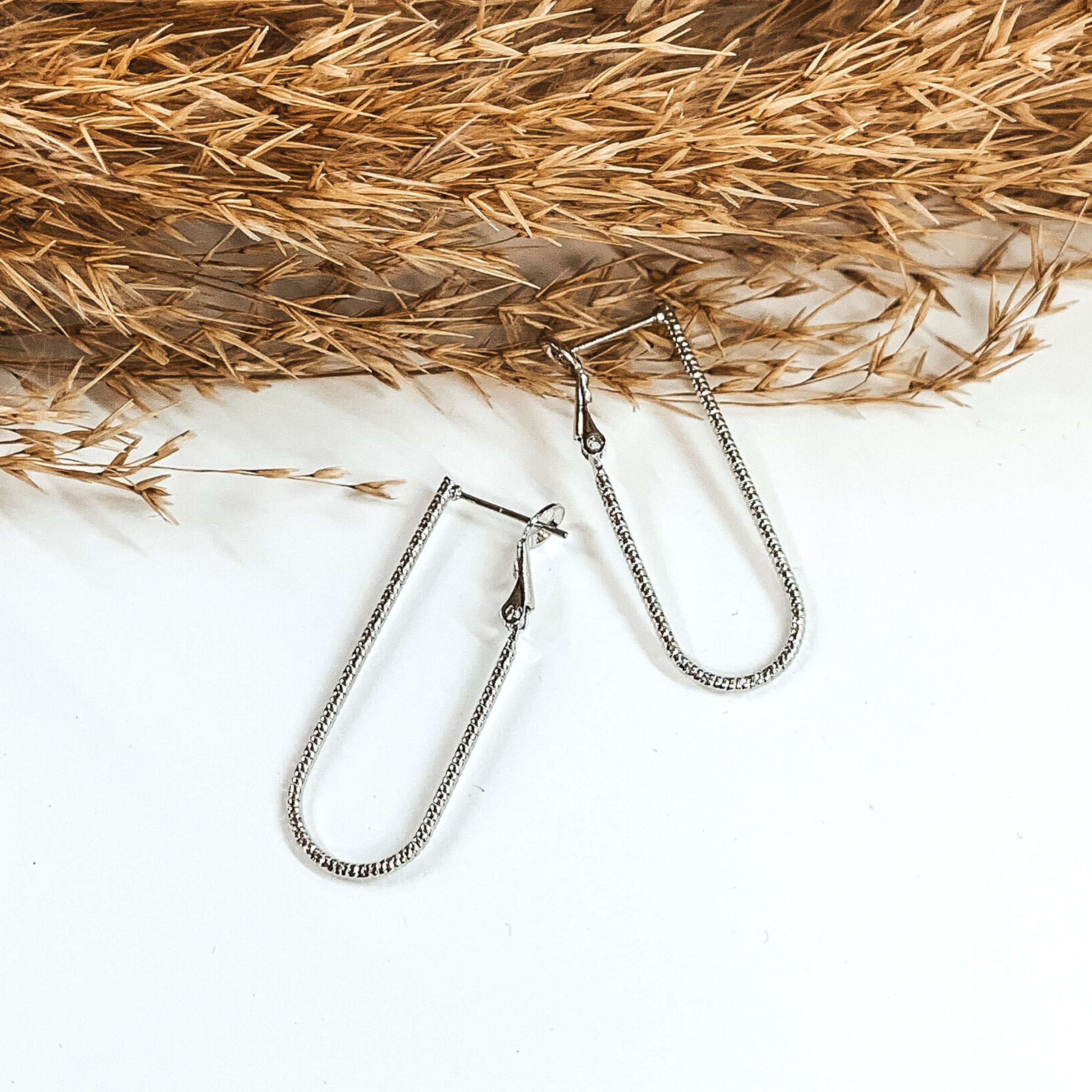 Silver u-shaped hoop earrings that have a striped pattern. These hoops are pictured on a white background and partially laying on some tan floral. 