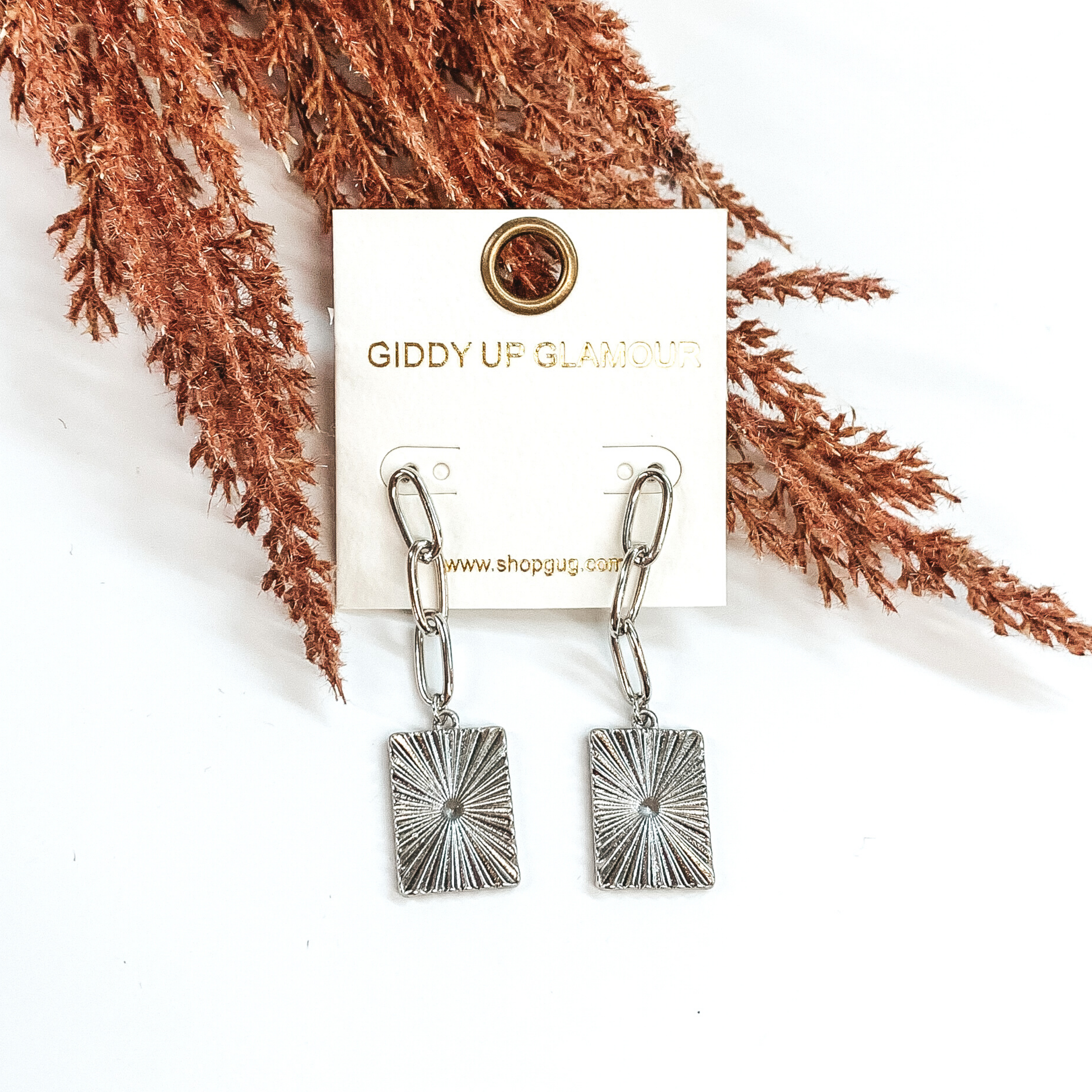 These are silver, chained, dangle earrings with a hanging rectangle pendant. The pendant has a starburst design. These earrings on partially laid on some brown floral on a white background. 