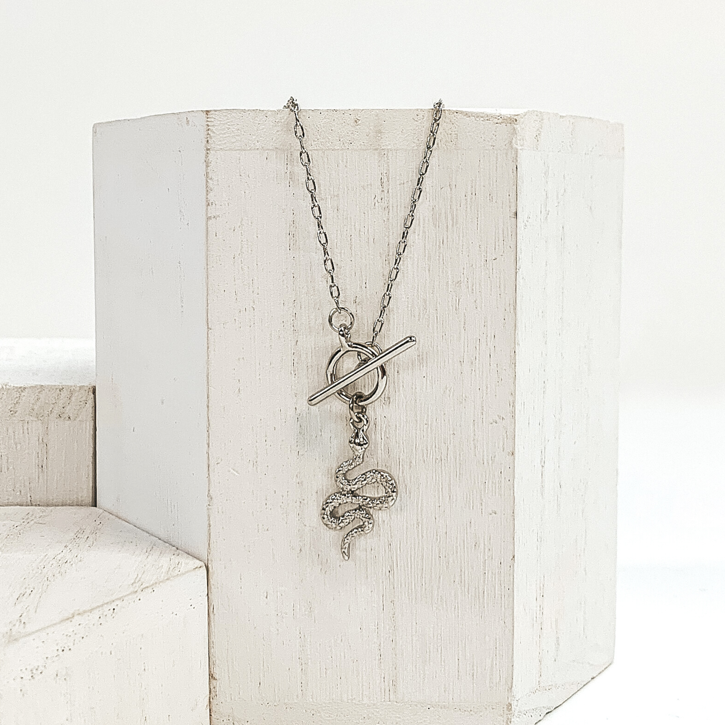This is a silver necklace that has a front toggle clasp and a silver snake charm. This necklace is pictured laying on a white block and on a white background with other white blocks around it.