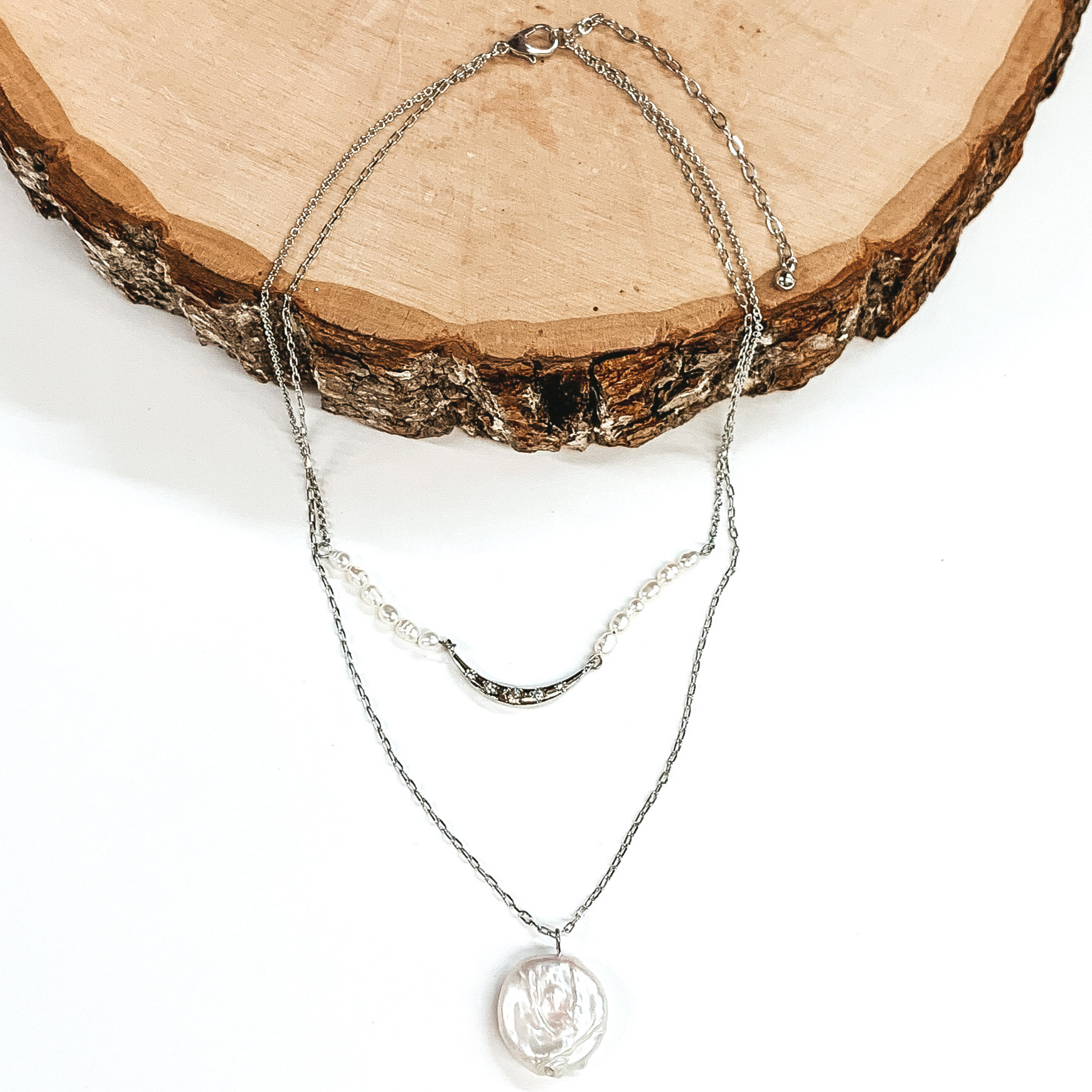 Silver douvle layered necklace. The shorter chain has a middle silver bar with white beads on either side of the bar. The longer strand has a single white pendant. This necklace is pictured laying partially on a piece of wood on a white background. 