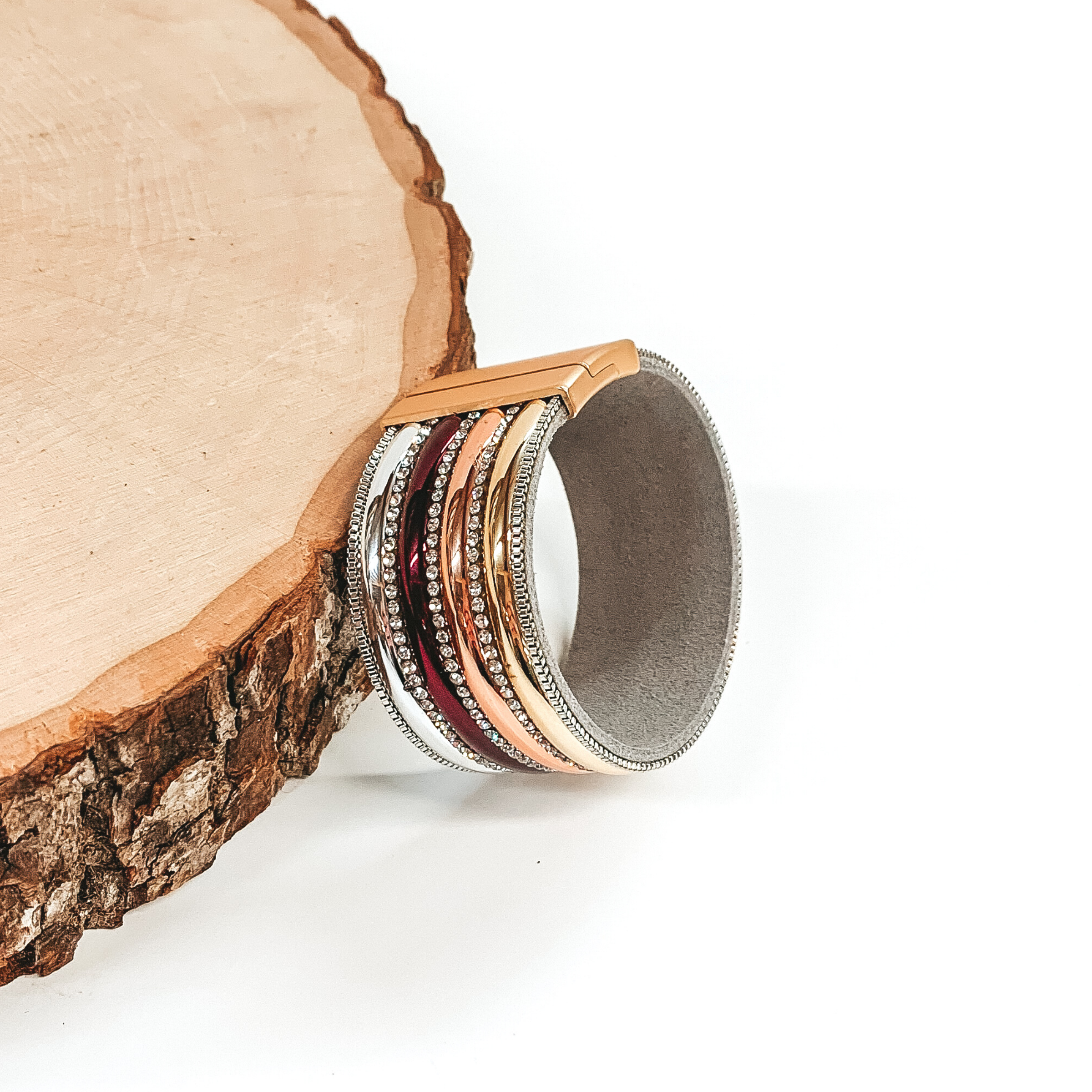 Wide band with rows of colors, including silver, maroon, rose gold, and gold, separated  by clear crystals. It has a gold clasp. This bracelet is pictured laying against a piece of wood on a white background. 
