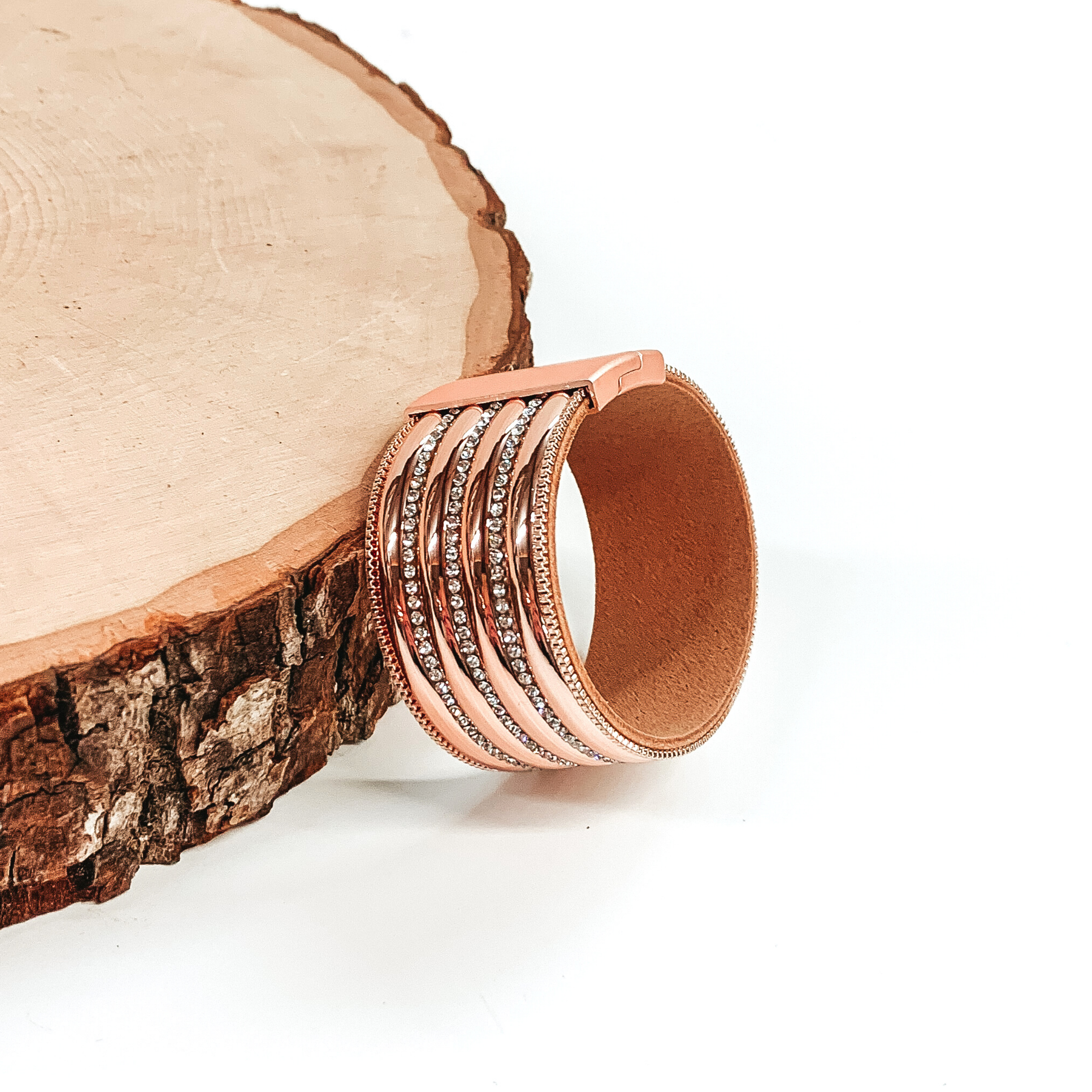 Wide band with rose gold colored rows separated by clear crystals. It has a rose gold clasp. This bracelet is pictured laying against a piece of wood on a white background.