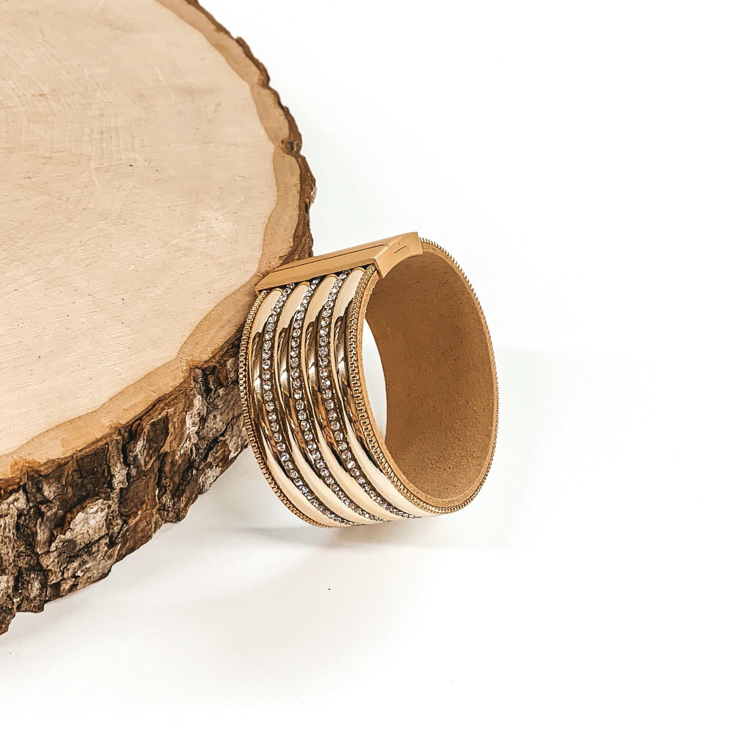 Wide band with gold colored rows separated by clear crystals. It has a gold clasp. This bracelet is pictured laying against a piece of wood on a white background.