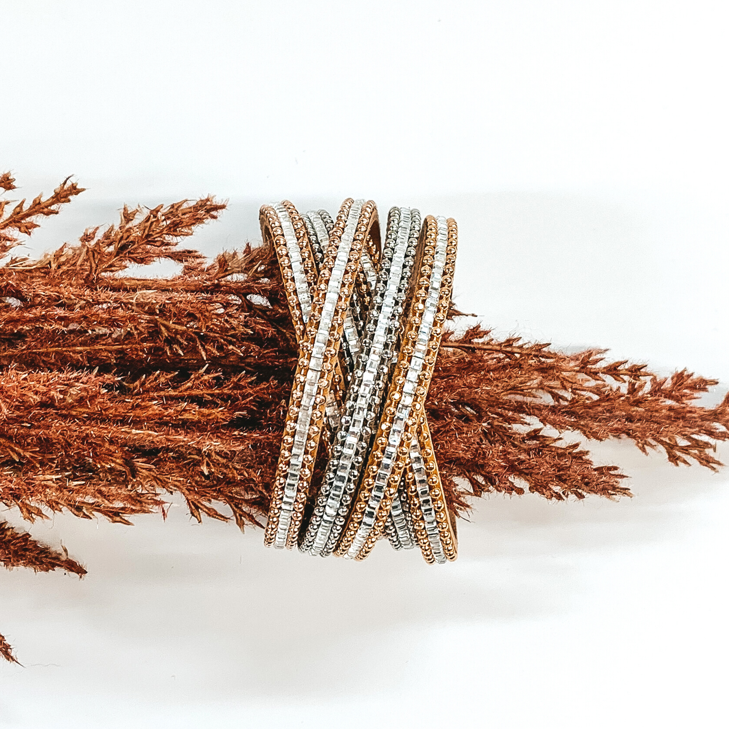 Gold and silver strands that has clear crystals in the middle of each strand. Two sections of three strands each are crossed over each other in the middle. This bracelet is pictured on a piece of brown floral on a white background. 