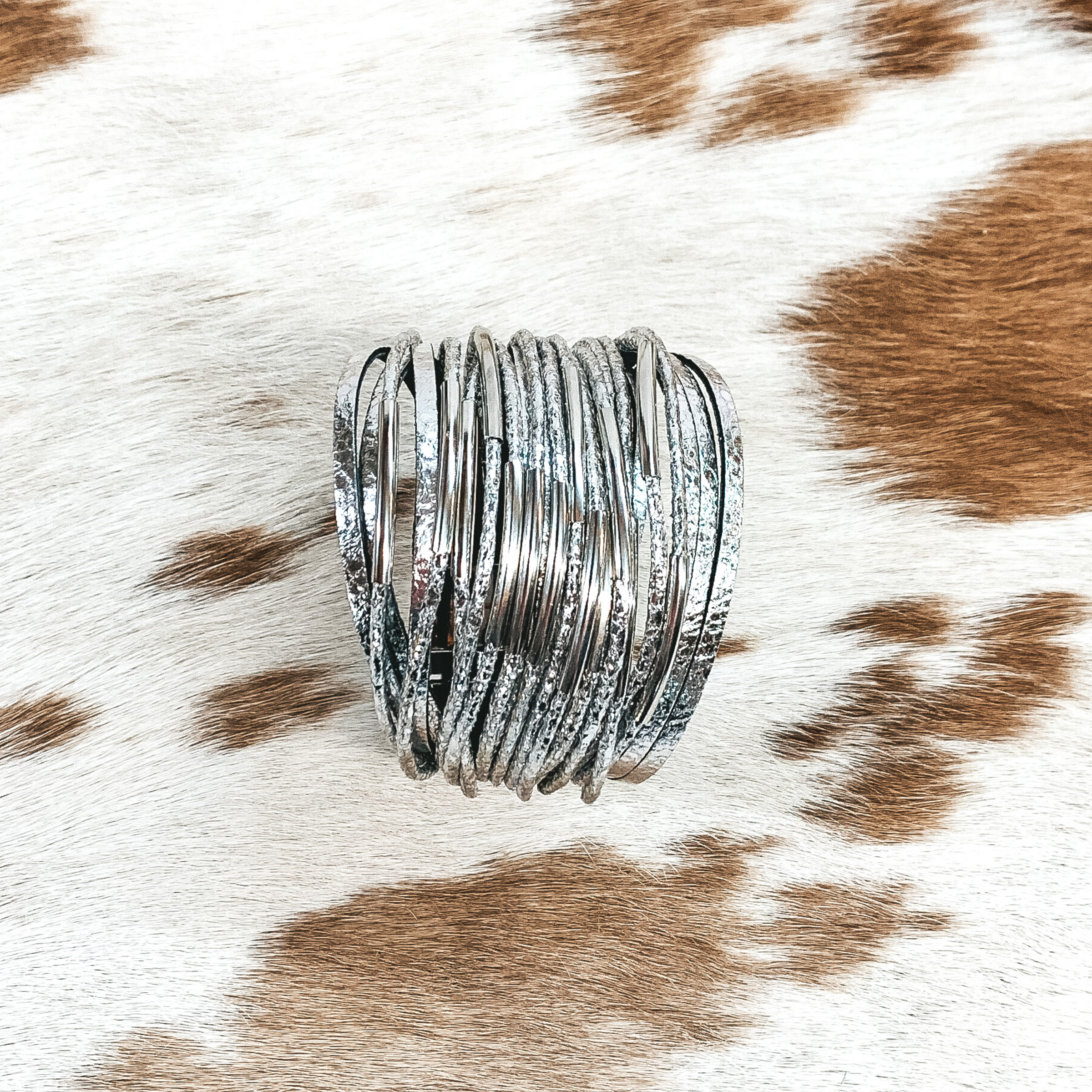 This bracelet has a lot of silver destressed strands with some of them having silver bar pendants. This bracelet is pictured on a white and brown cow print background.