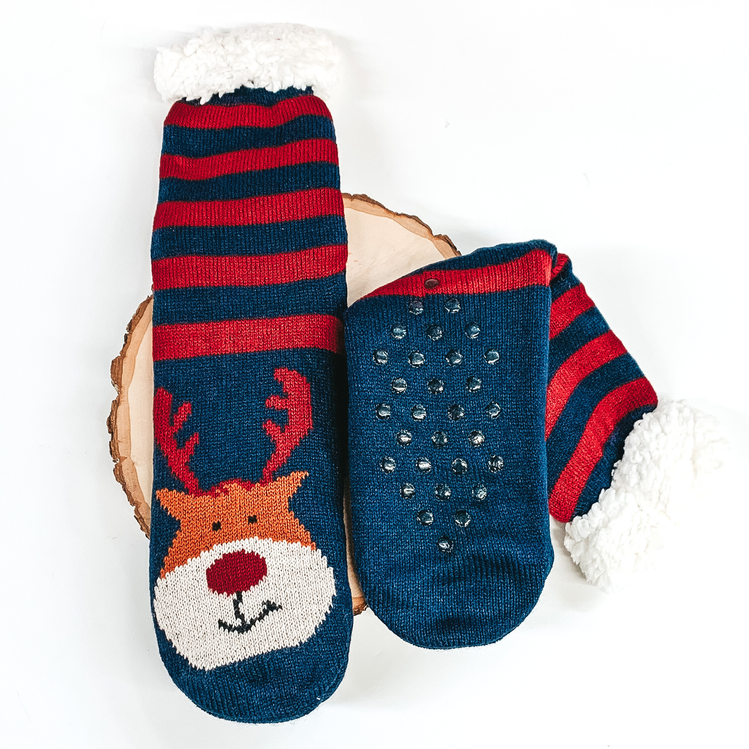 Navy socks that has Rudolph's face with red antlers towards the bottom of the socks. These socks also have stripes on the ankle parts, has rubber grips on the bottom, and white fluff on the top of the socks. These socks are pictured laying on a piece of wood on a white background.