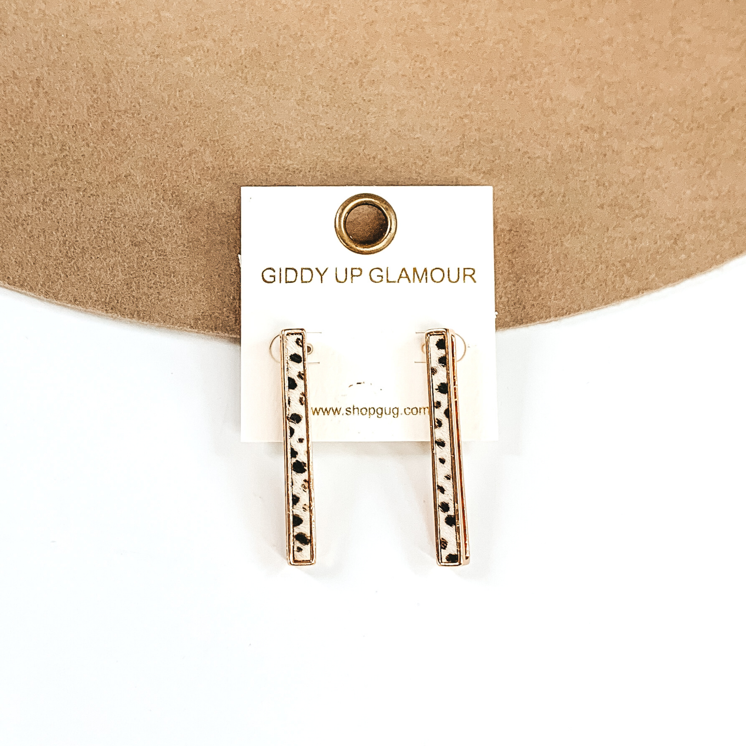 Gold rectangle bar earrings with a white dotted inlay on a white earrings holder. These earrings are pictured on a white and tan background.