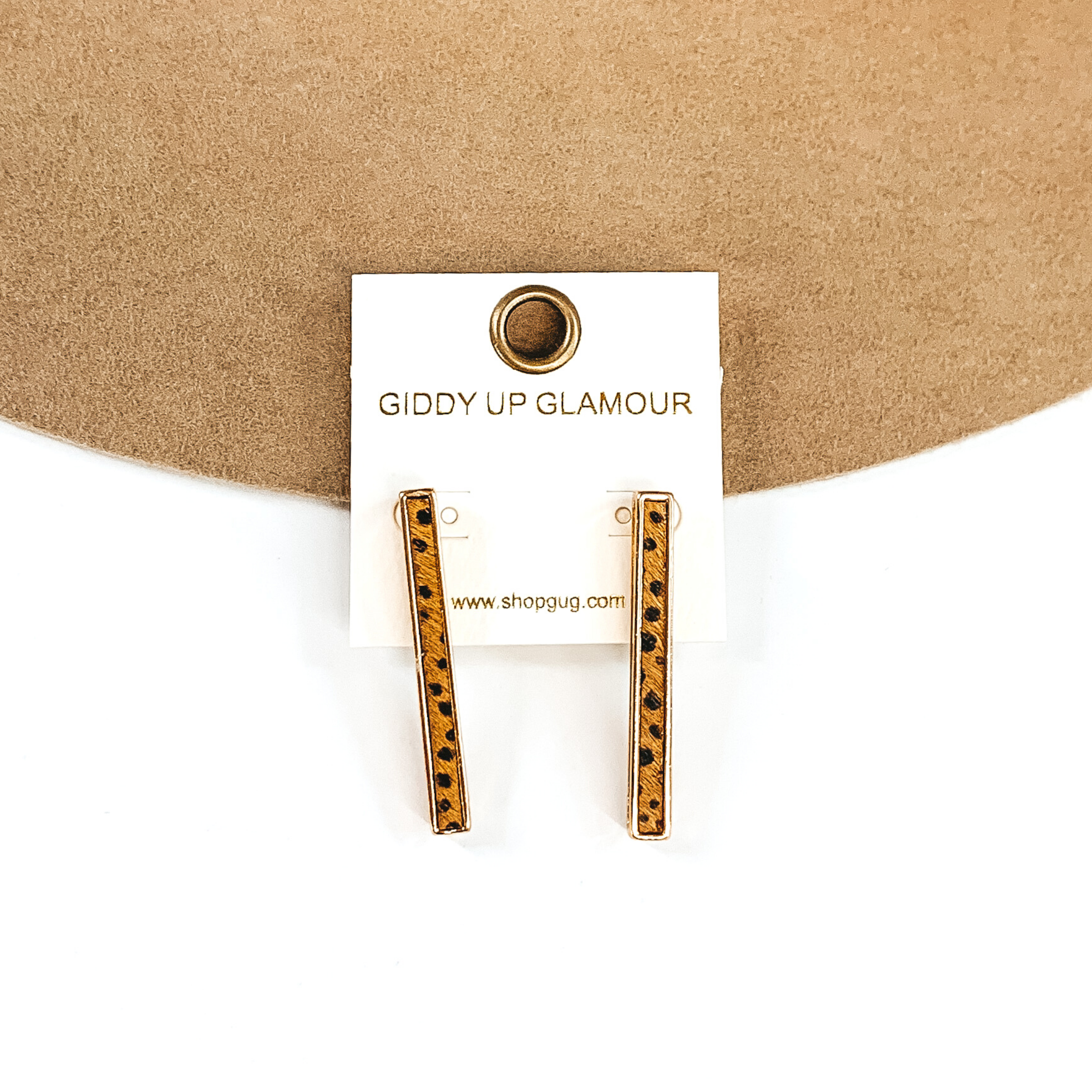 Gold rectangle bar earrings with a brown dotted inlay on a white earrings holder. These earrings are pictured on a white and tan background.