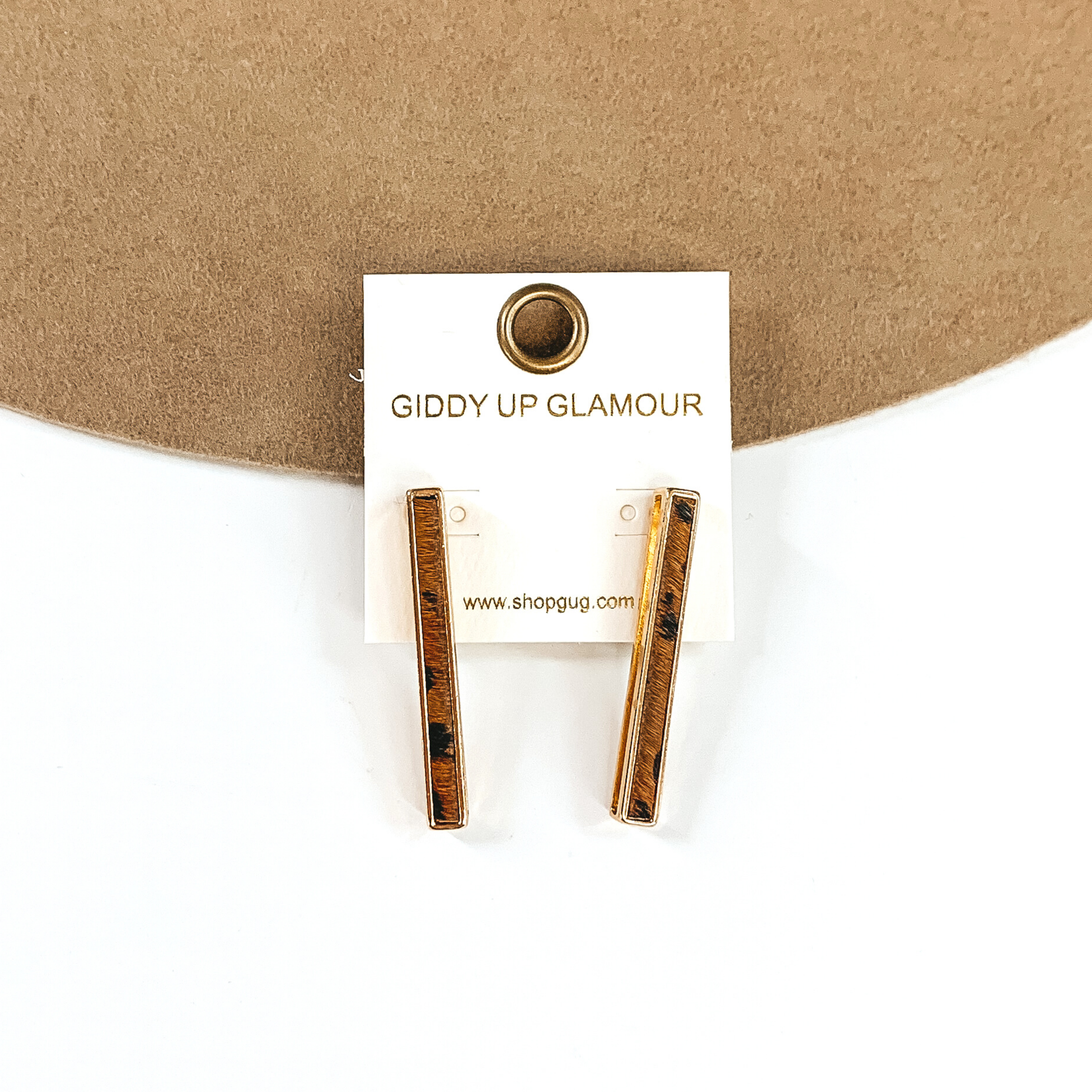 Gold rectangle bar earrings with a brown animal print inlay on a white earrings holder. These earrings are pictured on a white and tan background.