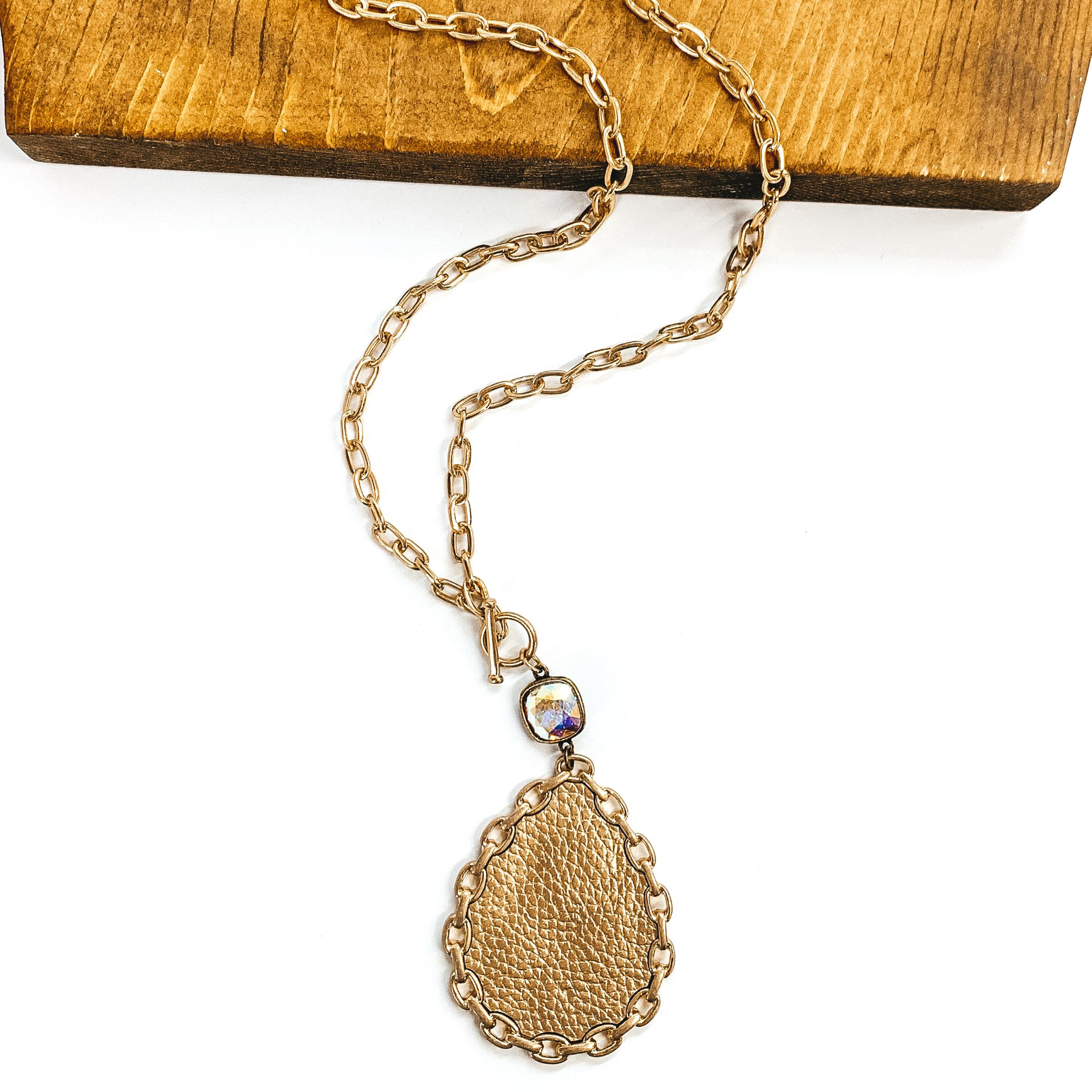 Gold chained necklace with front toggle clasp. This necklace includes a hanging AB cushion cut crystal with a bronze back, hanging from the toggle clasp. Hanging from the crystal was a teardrop pendant in gold with a gold chained outline. This necklace is pictured partially on a piece of dark wood and on a white background.