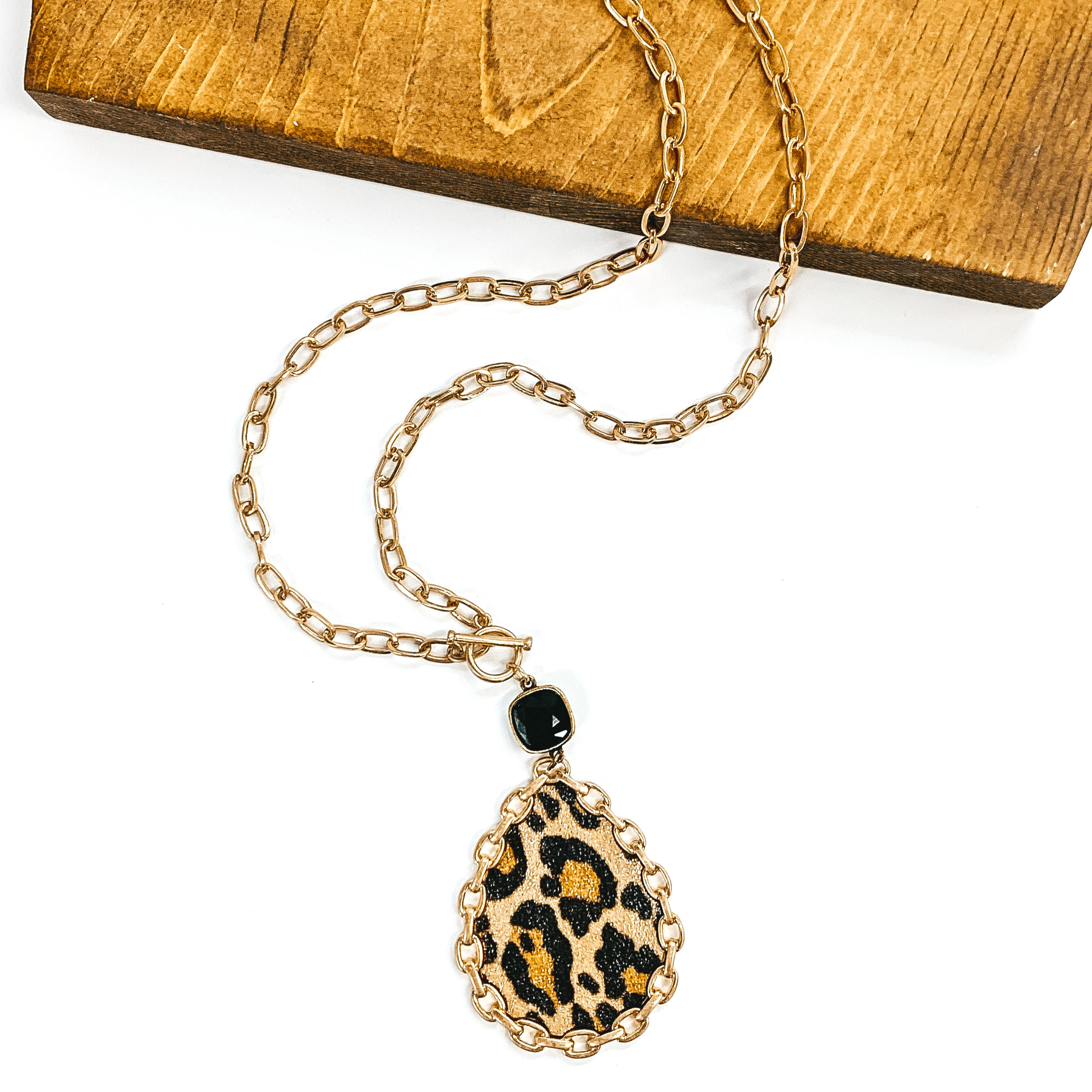 Gold chained necklace with front toggle clasp. This necklace includes a hanging black cushion cut crystal with a bronze back, hanging from the toggle clasp. Hanging from the crystal was a teardrop pendant in leopard print with a gold chained outline. This necklace is pictured partially on a piece of dark wood and on a white background.