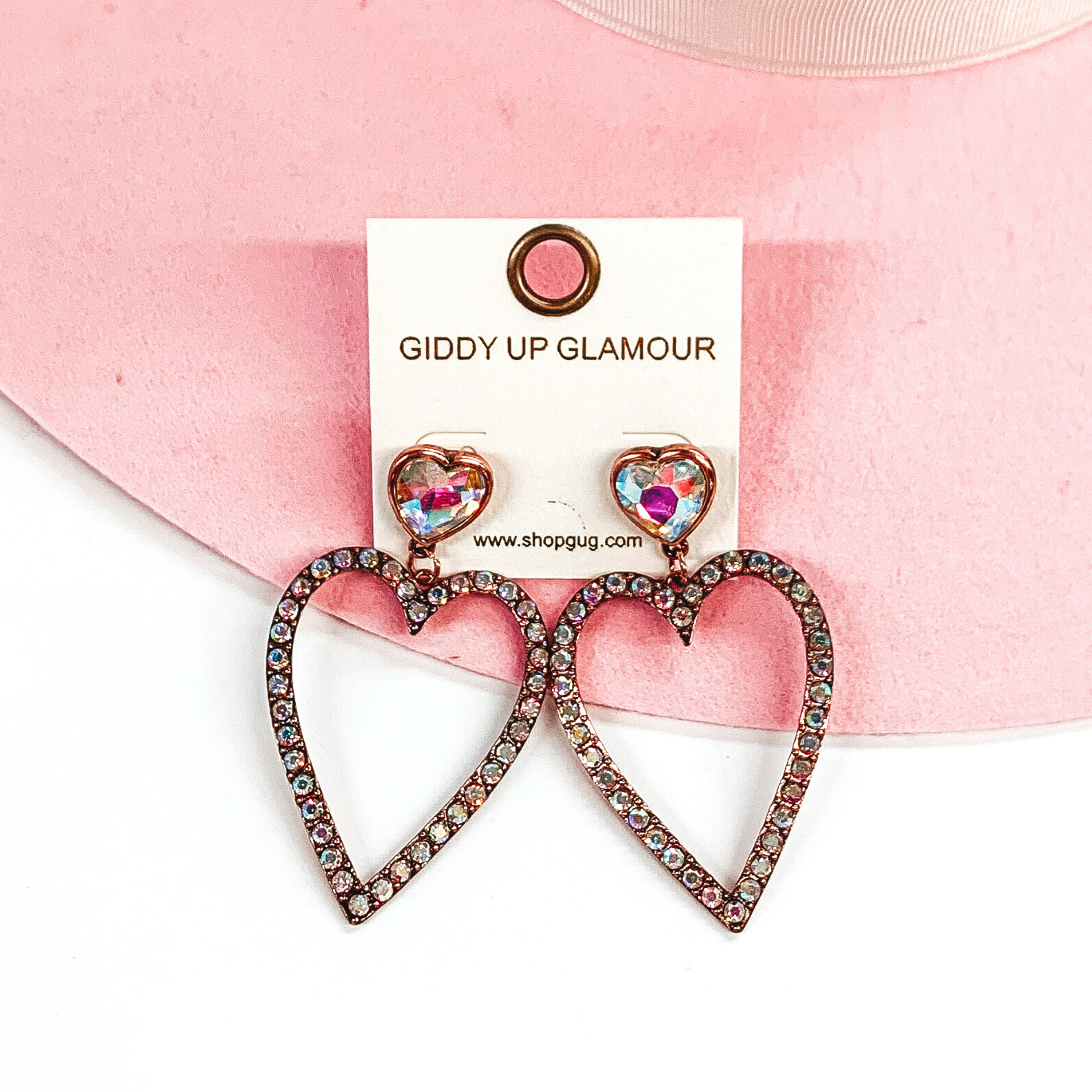 AB crystal heart shaped studs with a copper backing. The studs have a hanging heart outline pendant in copper with AB crystals. These earrings are pictured on a white and pink background. 