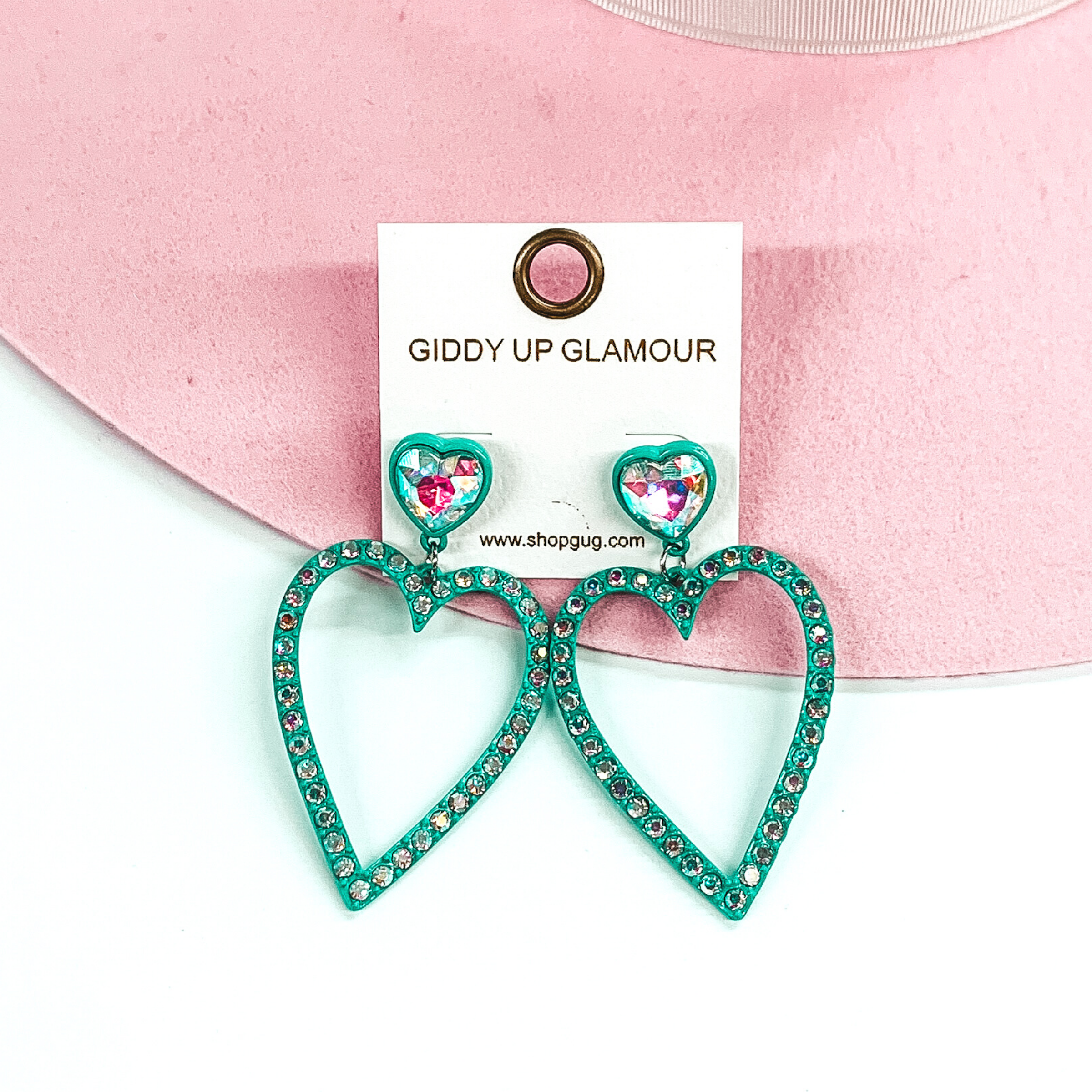 AB crystal heart shaped studs with a turquoise backing. The studs have a hanging heart outline pendant in turquoise with AB crystals. These earrings are pictured on a white and pink background. 