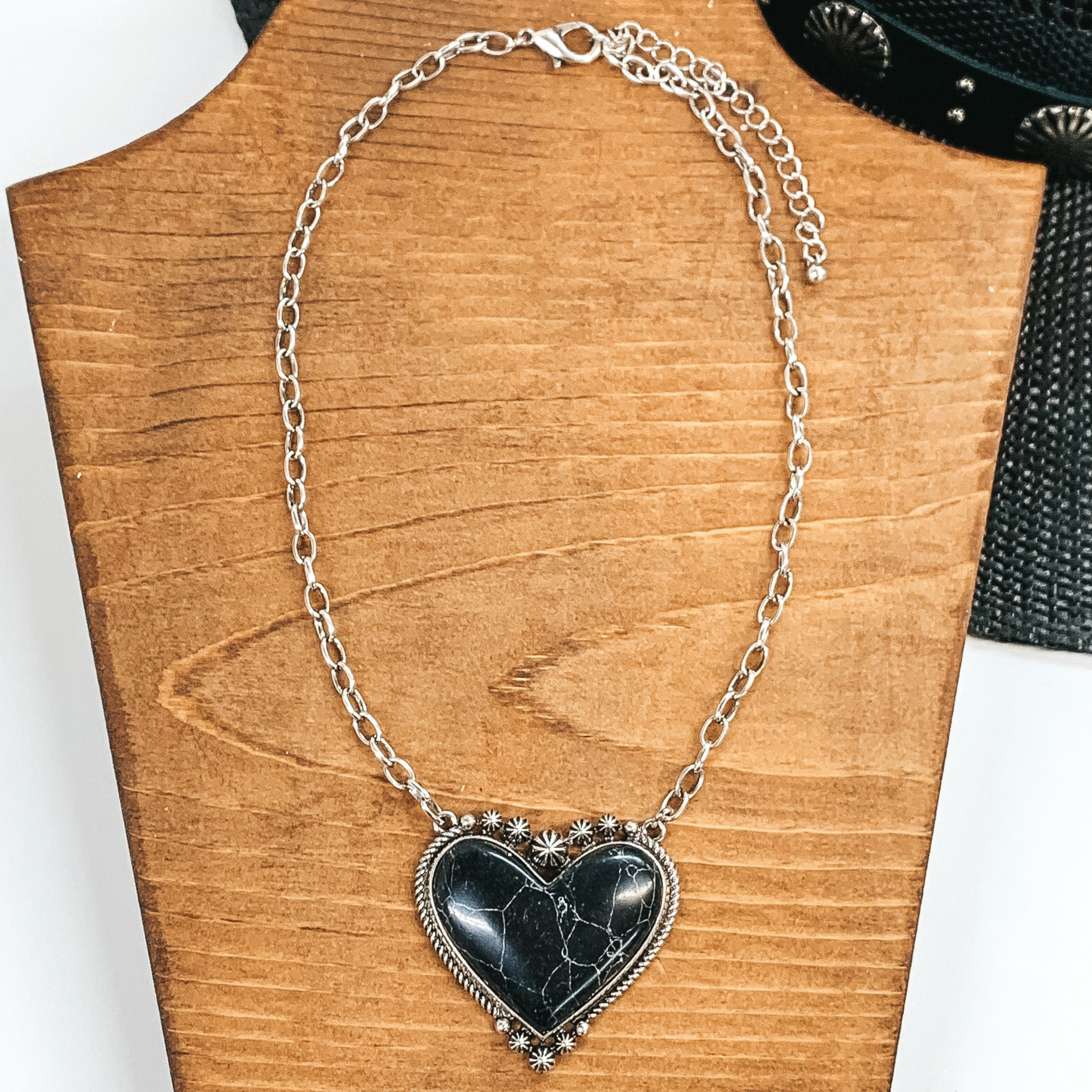 Silver chained necklace with heart pendant. The heart pendant is outlined in silver with a black stone. This necklace is pictured on a wood necklace holder on a white and black background. 