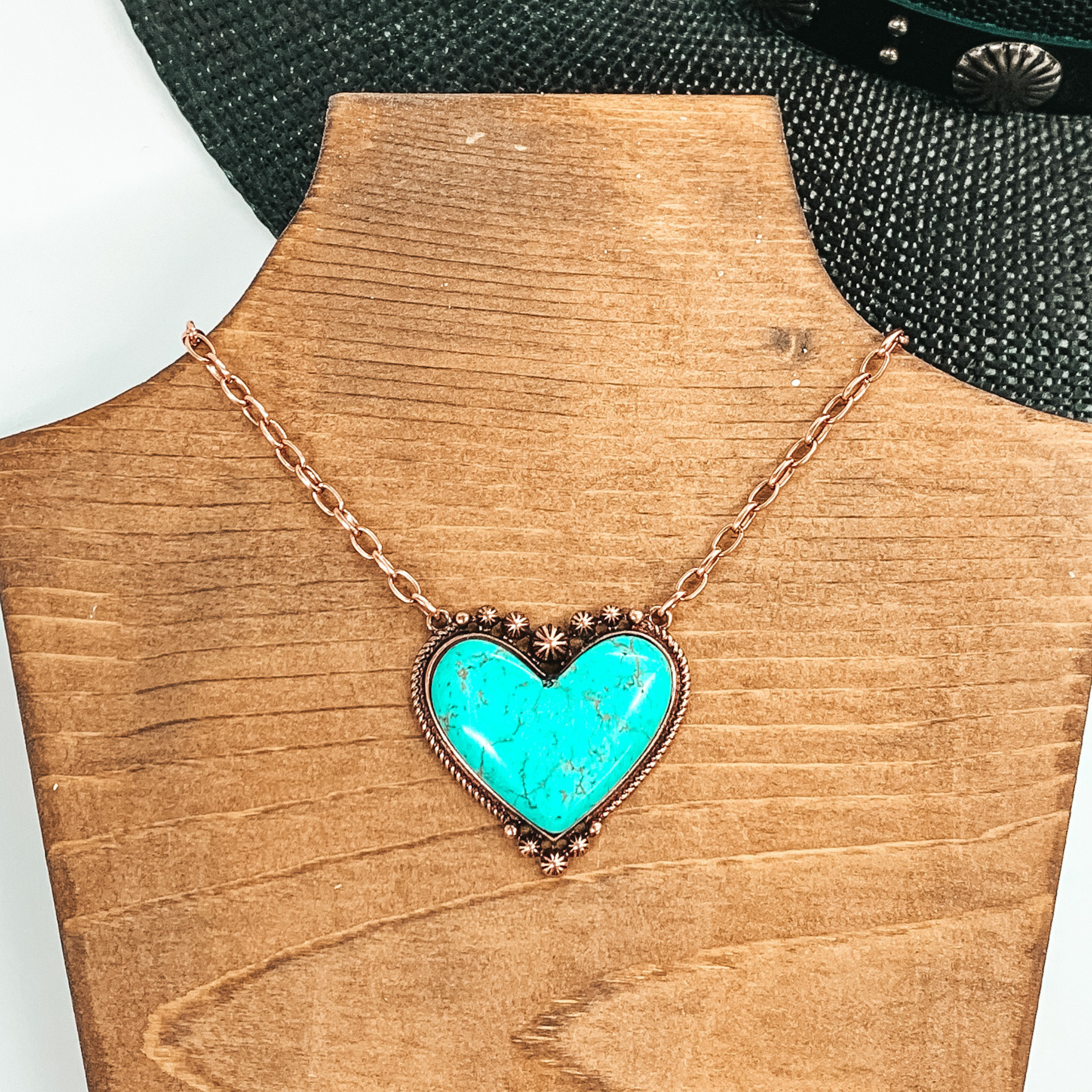 Copper chained necklace with a large turquoise, heart shaped stone pendant. This necklace is pictured laying on a brown necklace holder on a white and black background. 
