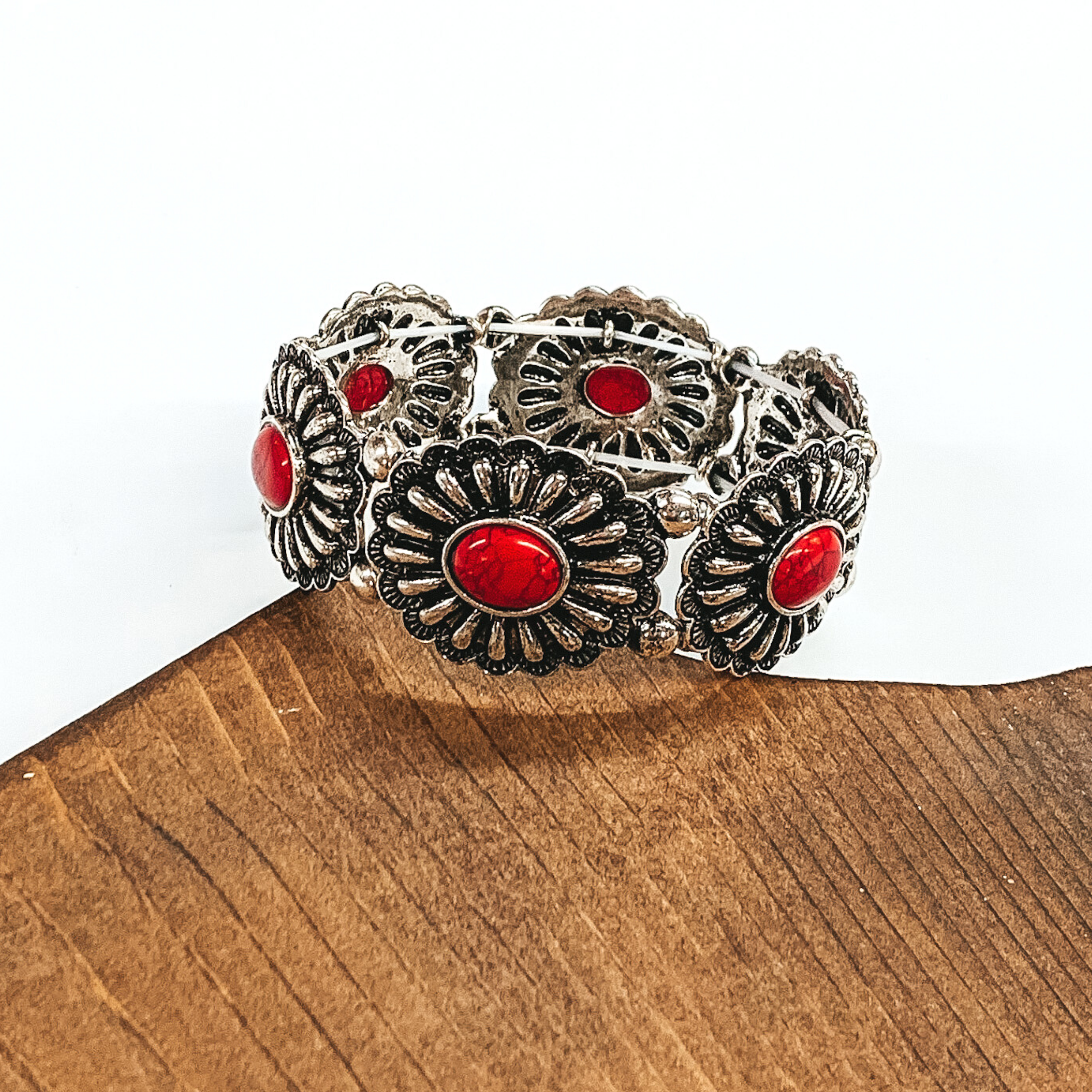 Silver, oval concho stretchy bracelet with oval center stones in red. This bracelet is pictured partially laying on a piece of wood and on a white background.