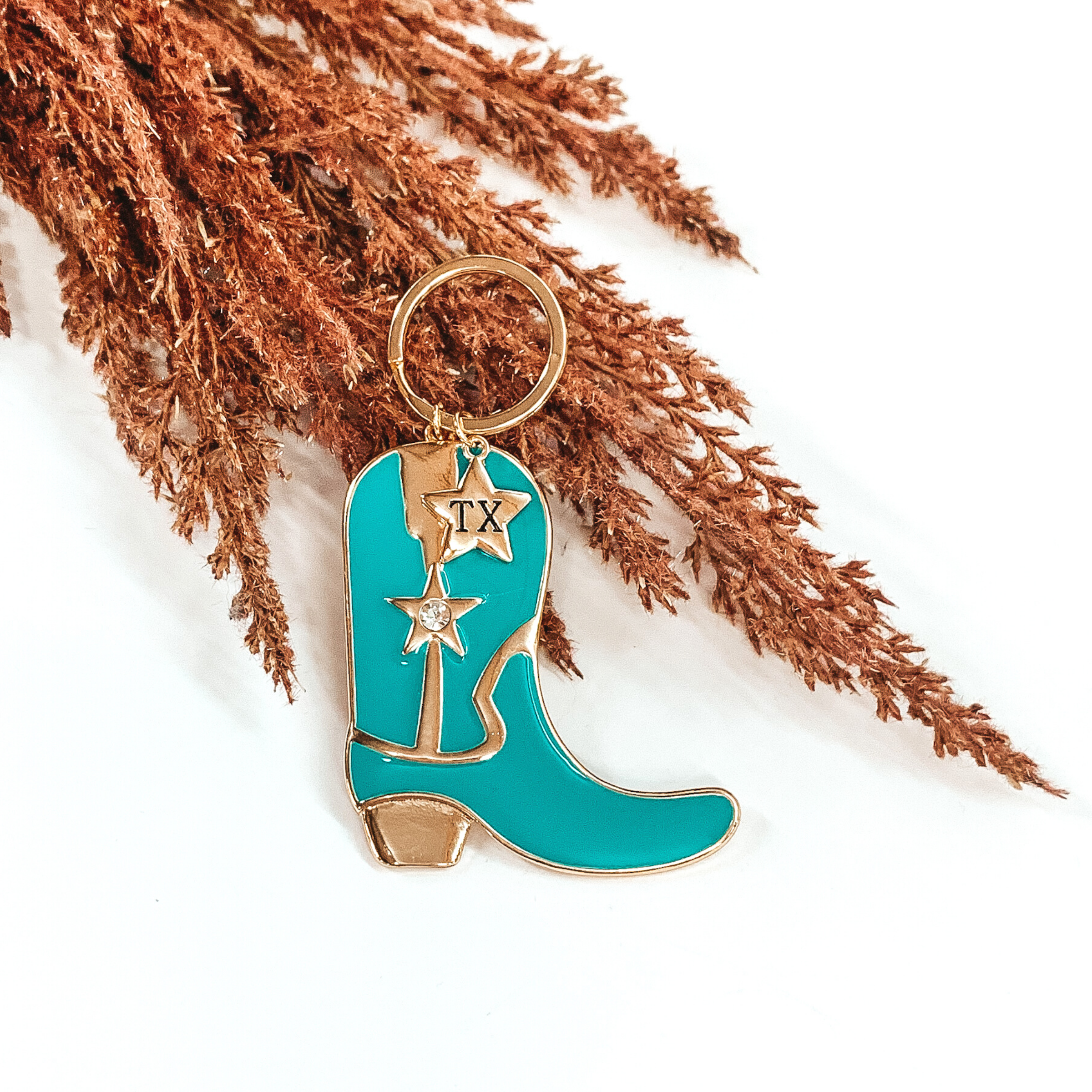 Turquoise cowboy boot key chain outline in gold. This key chain has a gold key ring and a gold star pendant with the letters "TX". This key chain is pictured laying on some brown floral on a white background. 