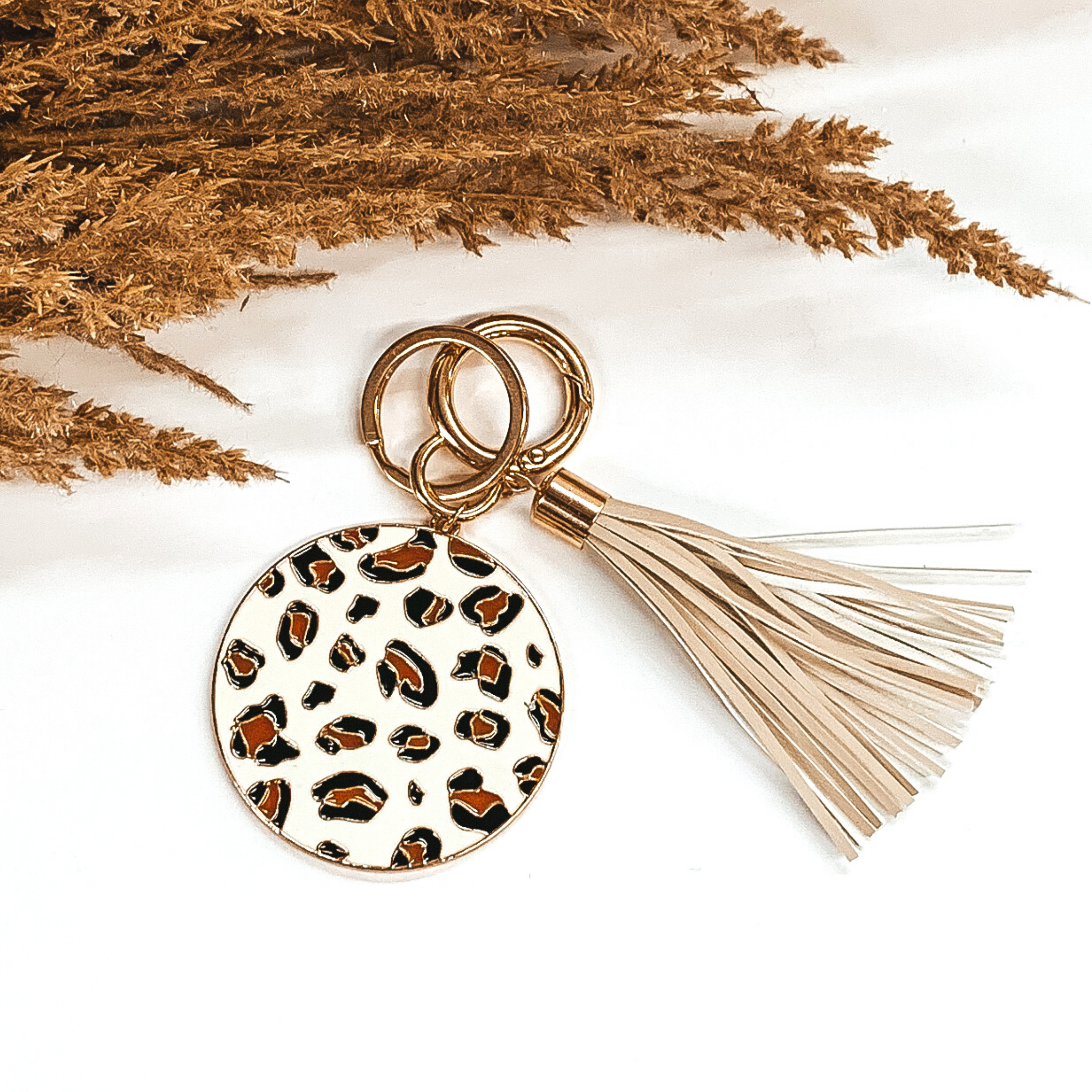 White circle key chain with a leopard print, two gold key rings, and an ivory tassel. This key chain is pictured laying on a white background with some brown floral at the top of the picture. 