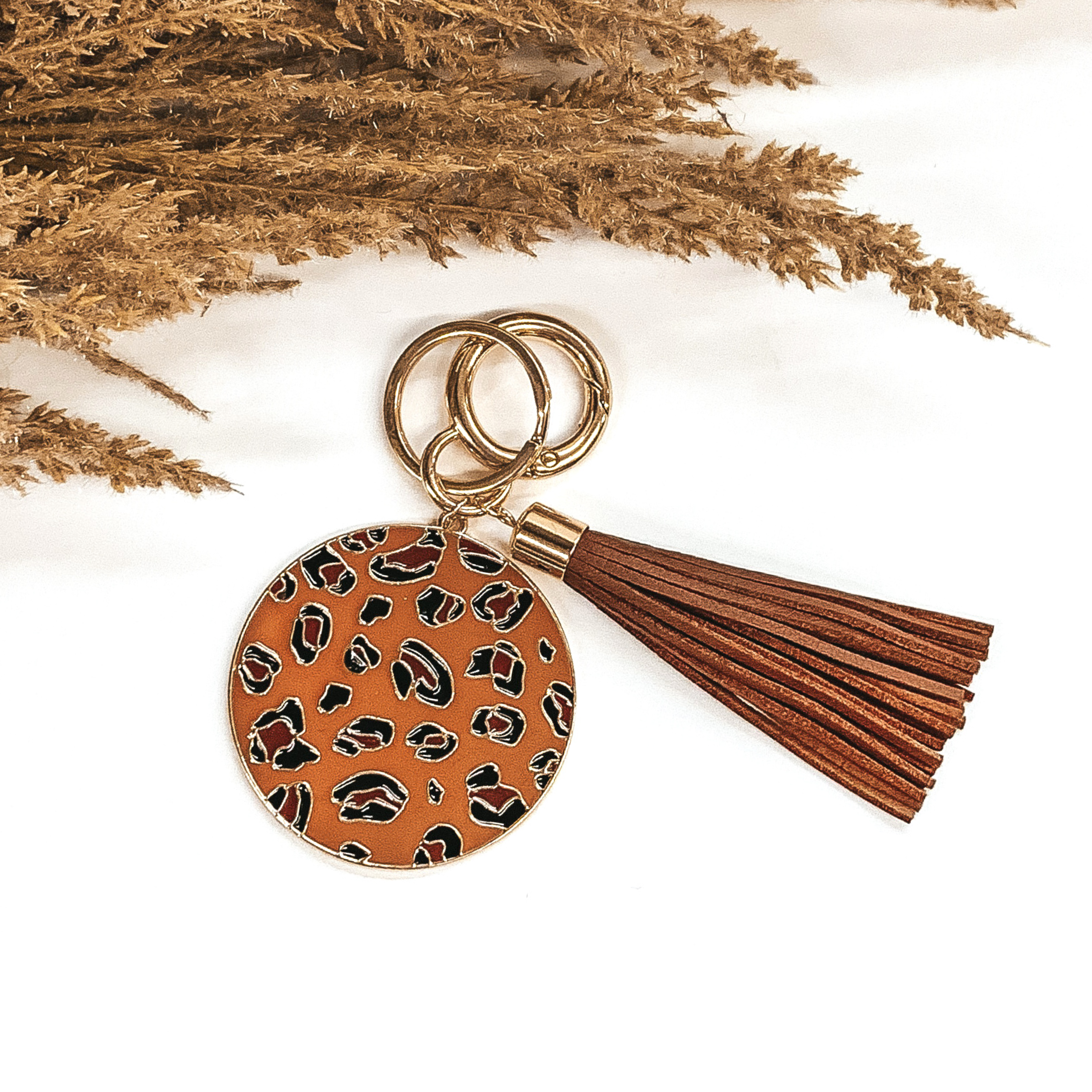 Dark tan circle key chain with a leopard print, two gold key rings, and a brown tassel. This key chain is pictured laying on a white background with some brown floral at the top of the picture.