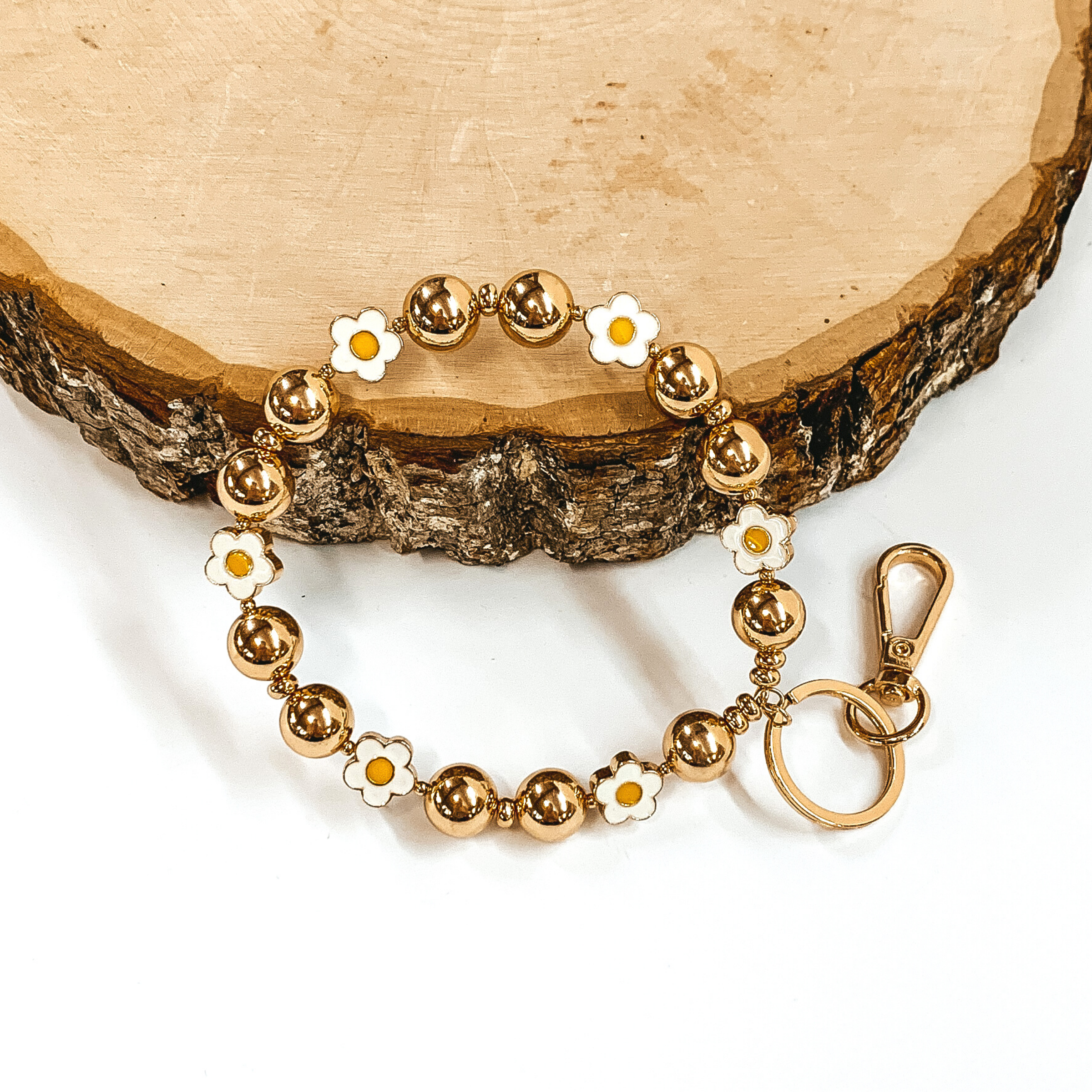 Gold beaded bracelet with white flower spacers. There is a gold key ring and clasp on one end of the bangle. It is pictured laying against a piece of wood on a white background.