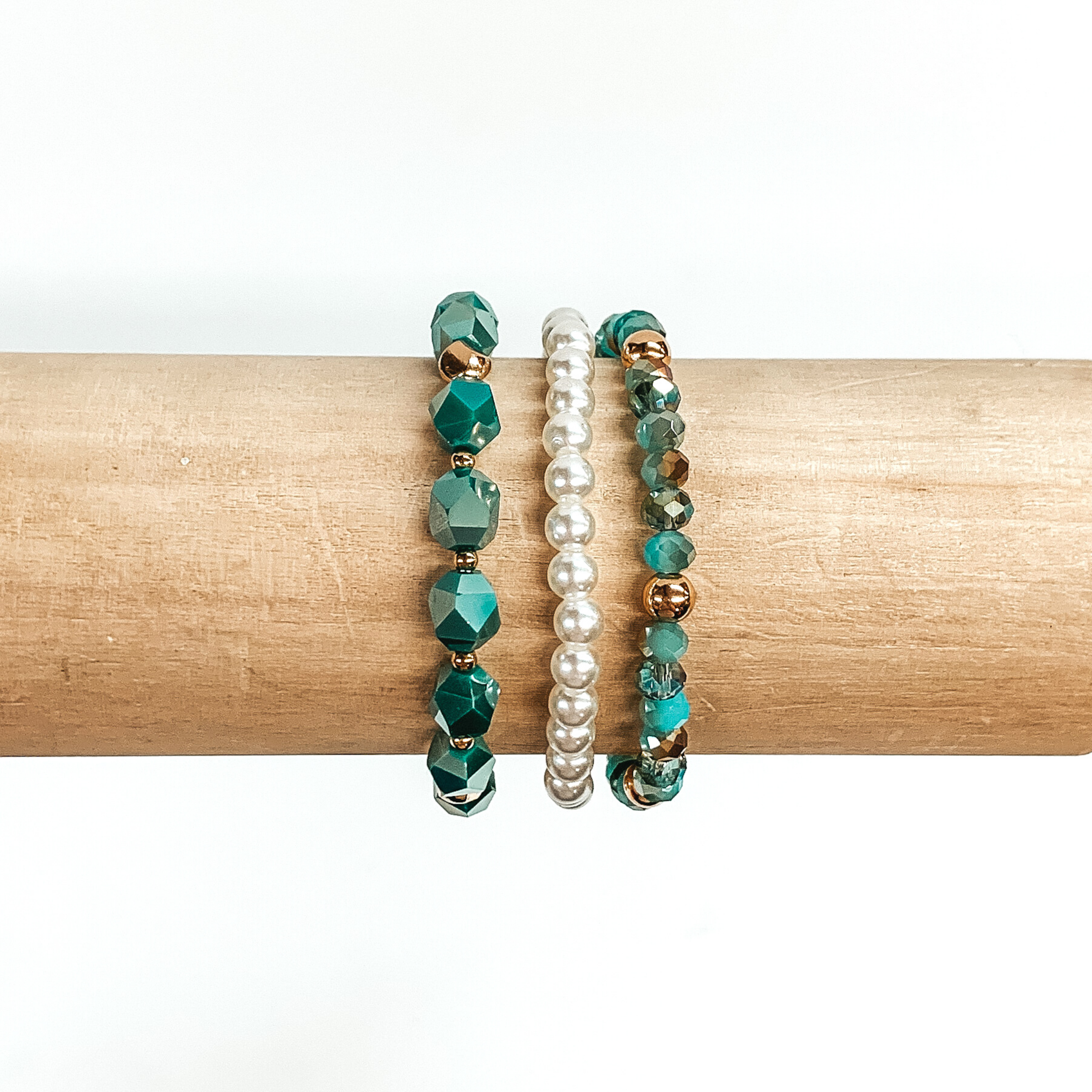 Beaded bracelet set with three different bracelets. One bracelet is white pearl beads only. The next one has big teal crystal beads with gold spacers. The last braclet has a mixture of tan and teal crystal beads with gold spacers. These bracelets are pictured on a wood bracelet holder on a white background.