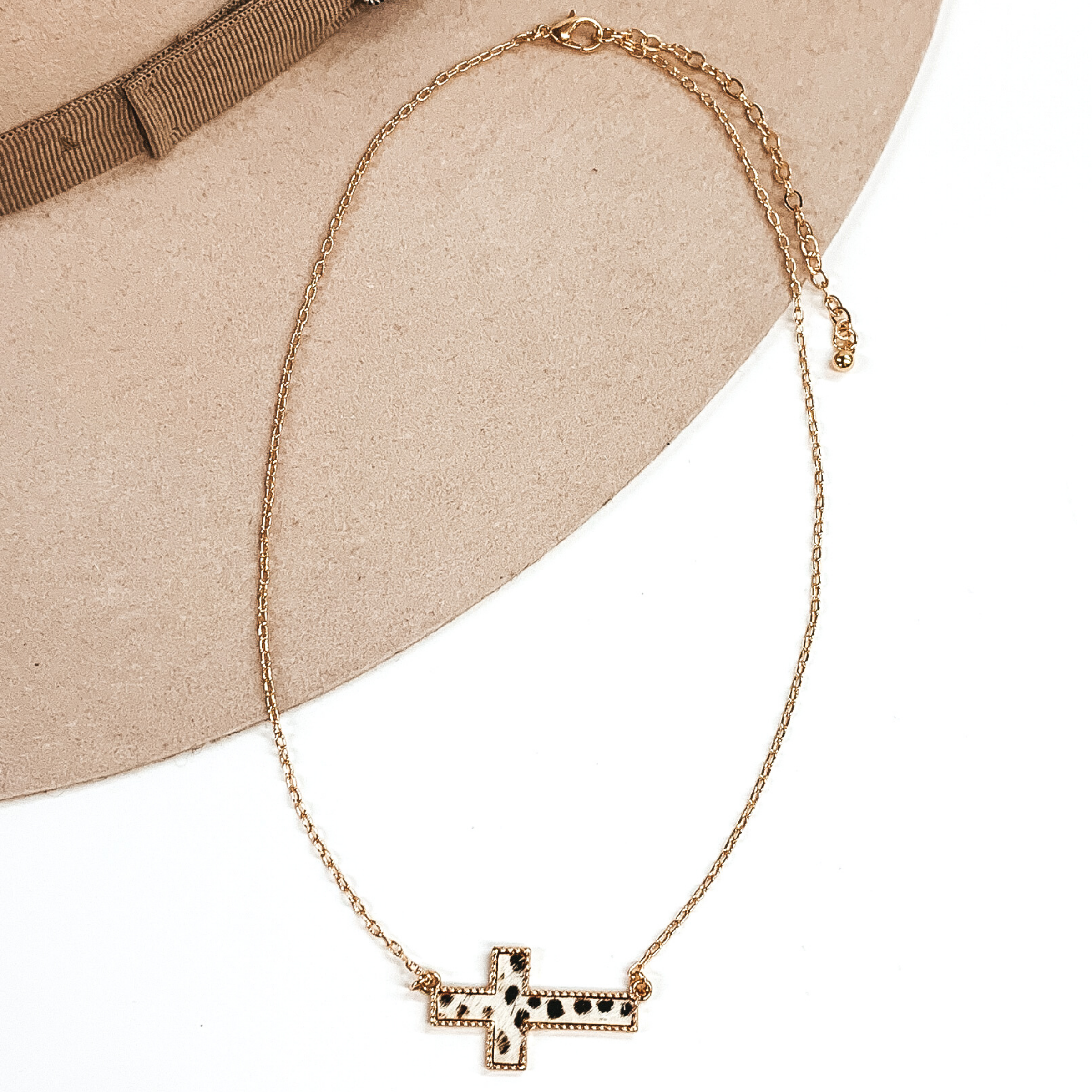 Gold paperclip chain with a cross pendant. The pendant has a white hide inlay with black dots. this necklace is pictured on a white and beige background. 