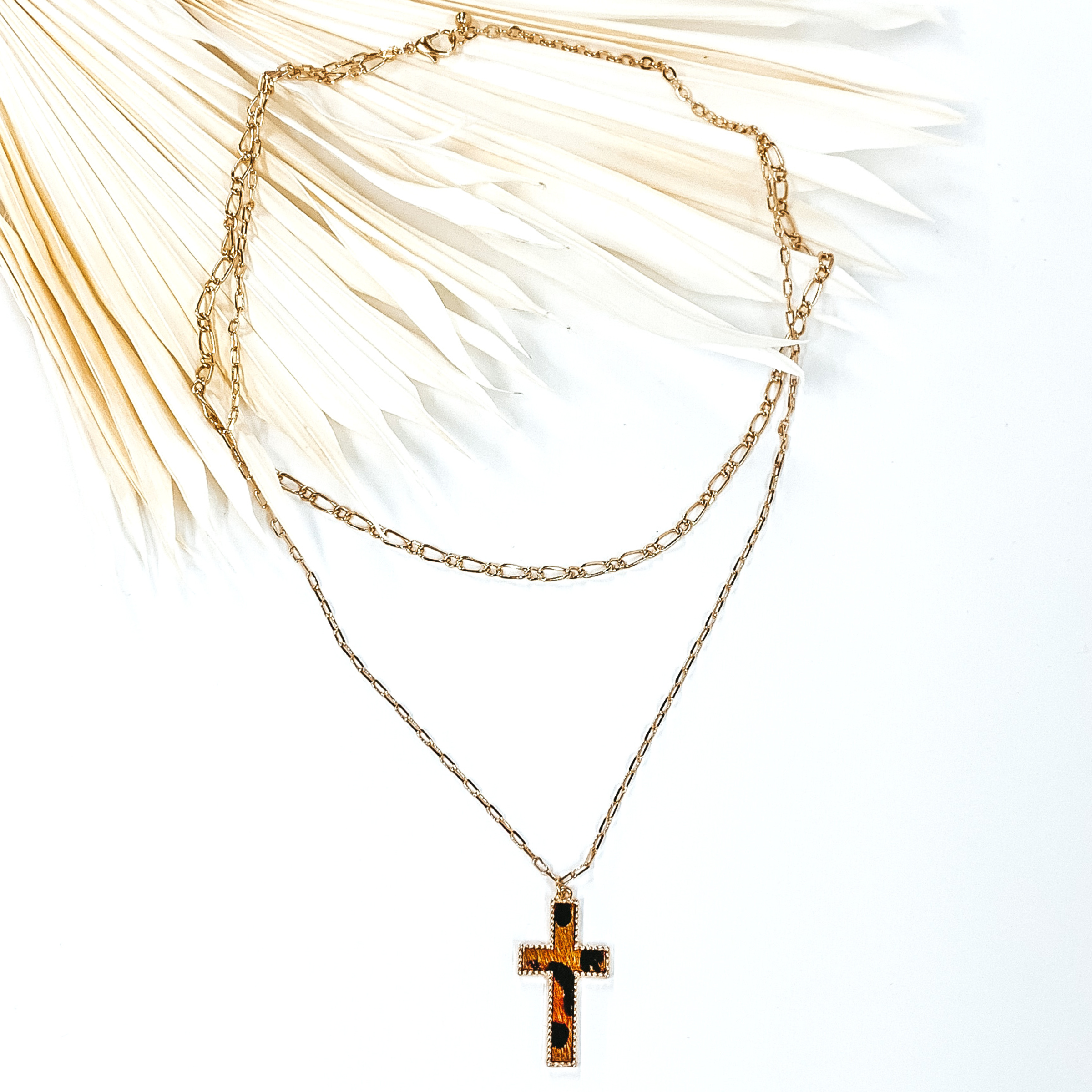 Gold paperclip doubled chain with a cross pendant. The pendant has a brown hide inlay with an animal print. This necklace is pictured laying on cream leaf on a white background.