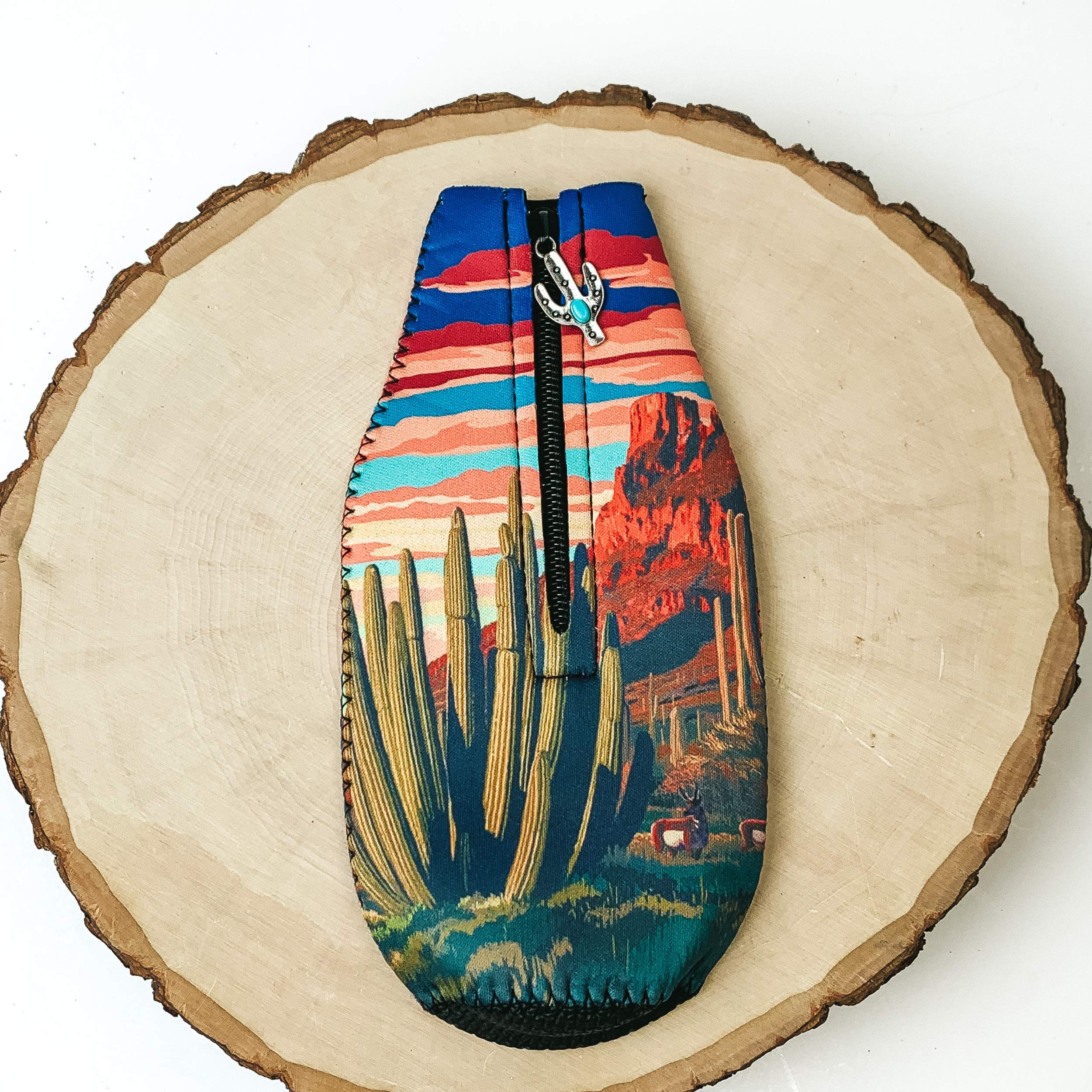 This Cactus Landscape Zip Up Koozie with Multicolored Sky with Cactus Charm is pictured on a peice of wood, with a white background. 