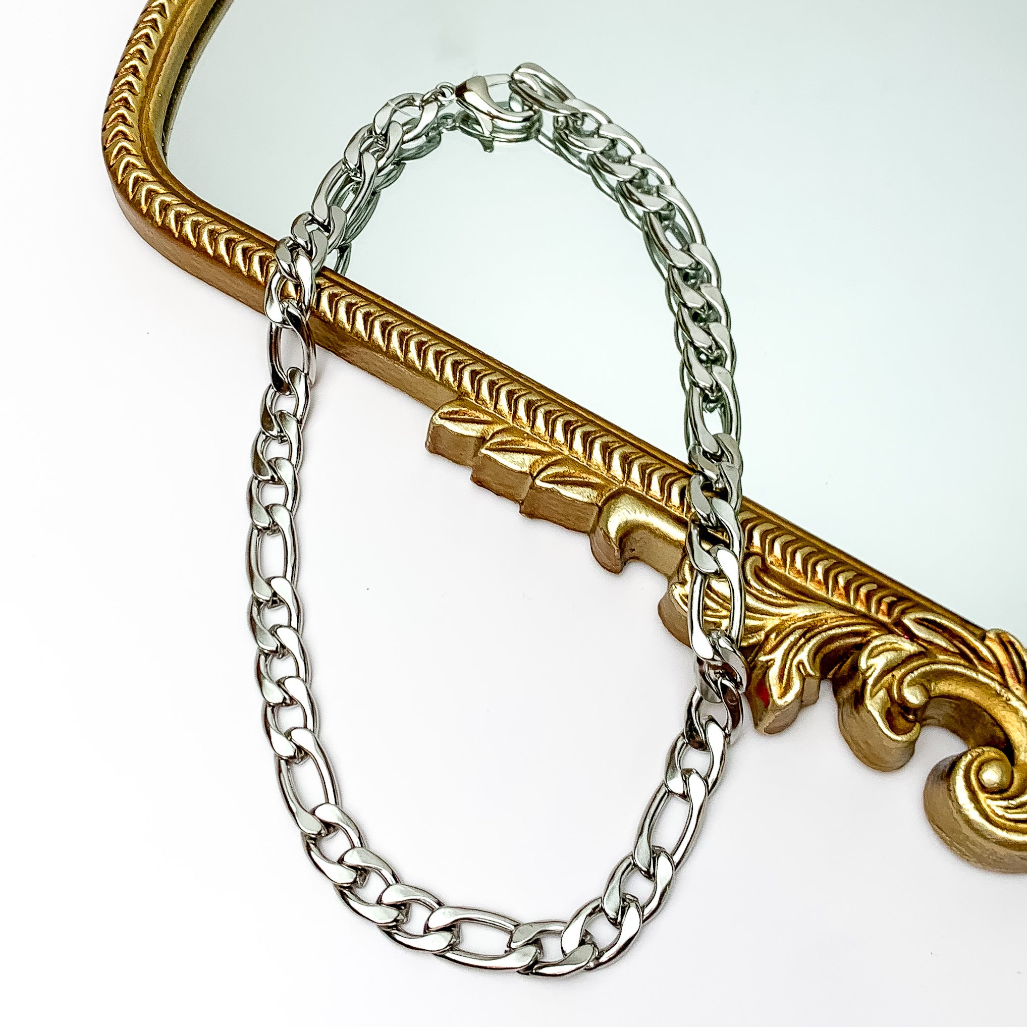 Pictured is a silver, curb chain necklace. This necklace is pictured partially laying on a gold mirror on a white background.  