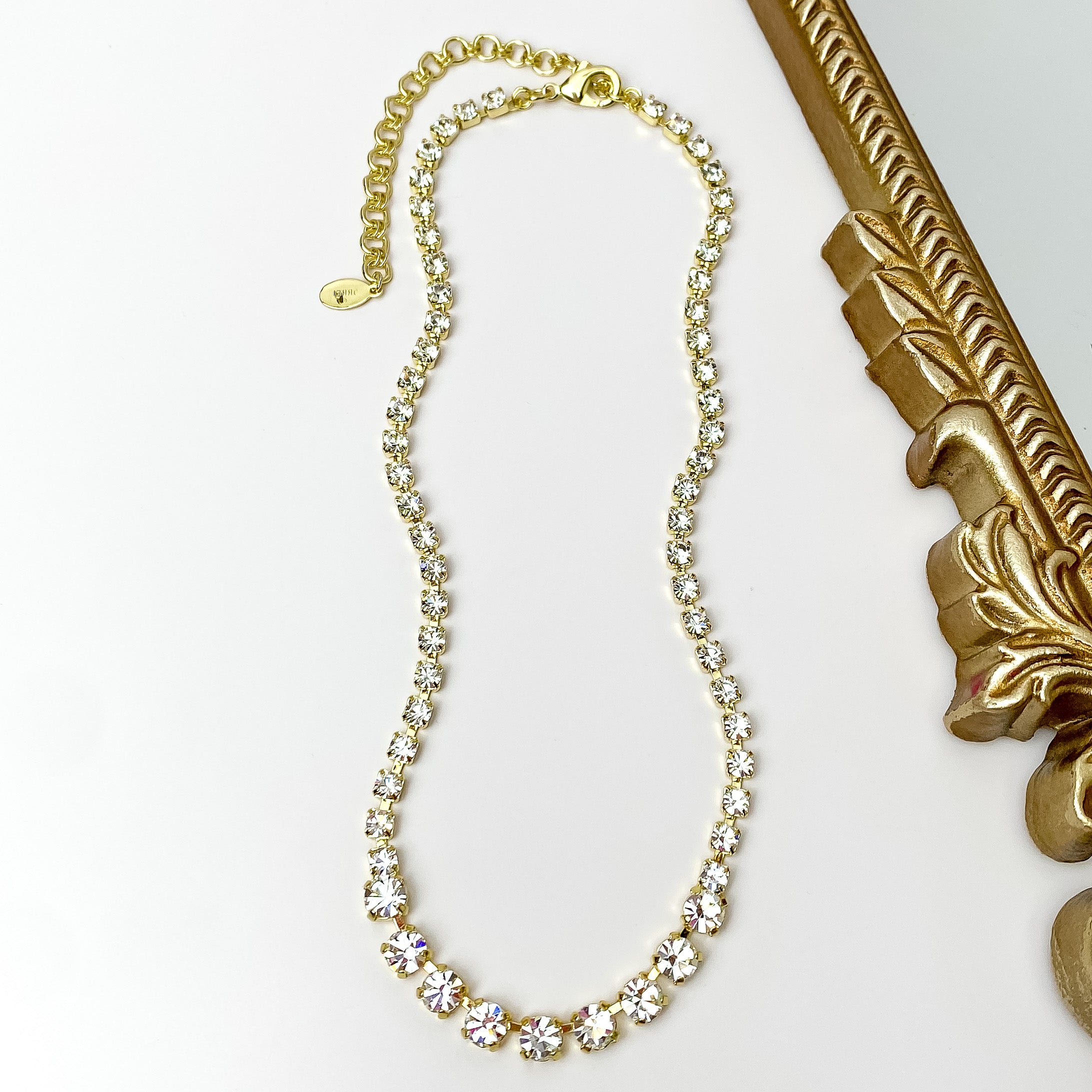 Pictured is a gold necklace with clear crystals. This necklace is pictured on a white background with a gold mirror on the right side.