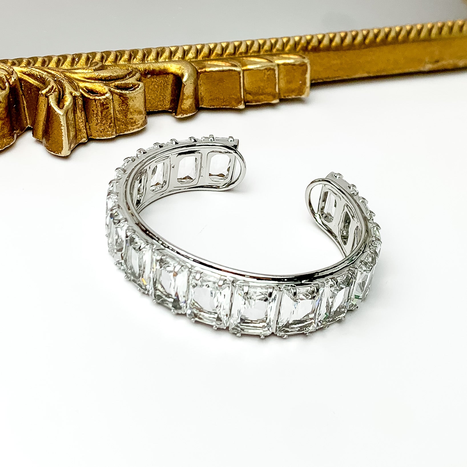 Silver bangle with a clear, rectangle crystal inlay. This bracelet is pictured on a white background with a gold mirror at the top of the picture.  