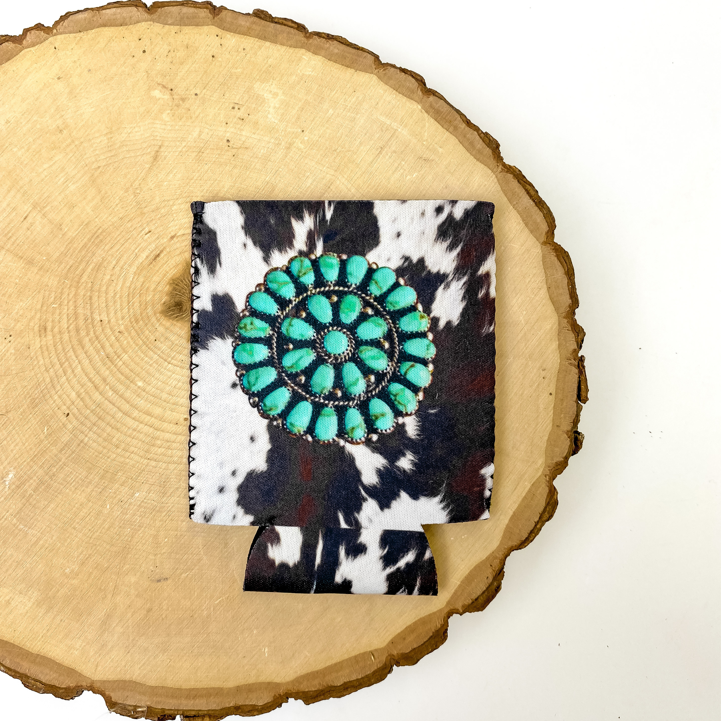 Cow print design with a turquoise design in the center of koozie. This koozie is pictured on a piece of wood on a white background.