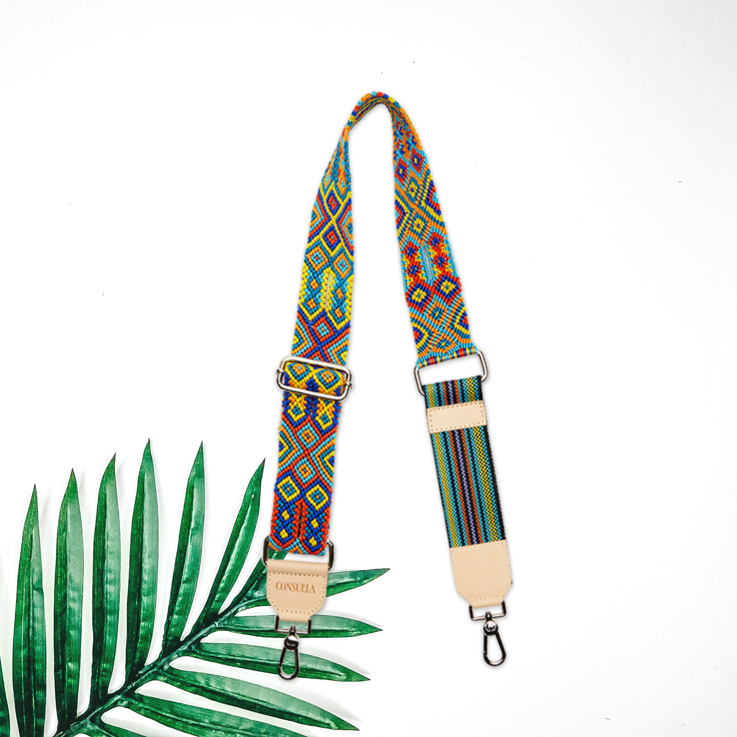 Centered in the picture is a woven crossbody purse strap. To the left of the strap is a palm leaf, all on a white background. 
