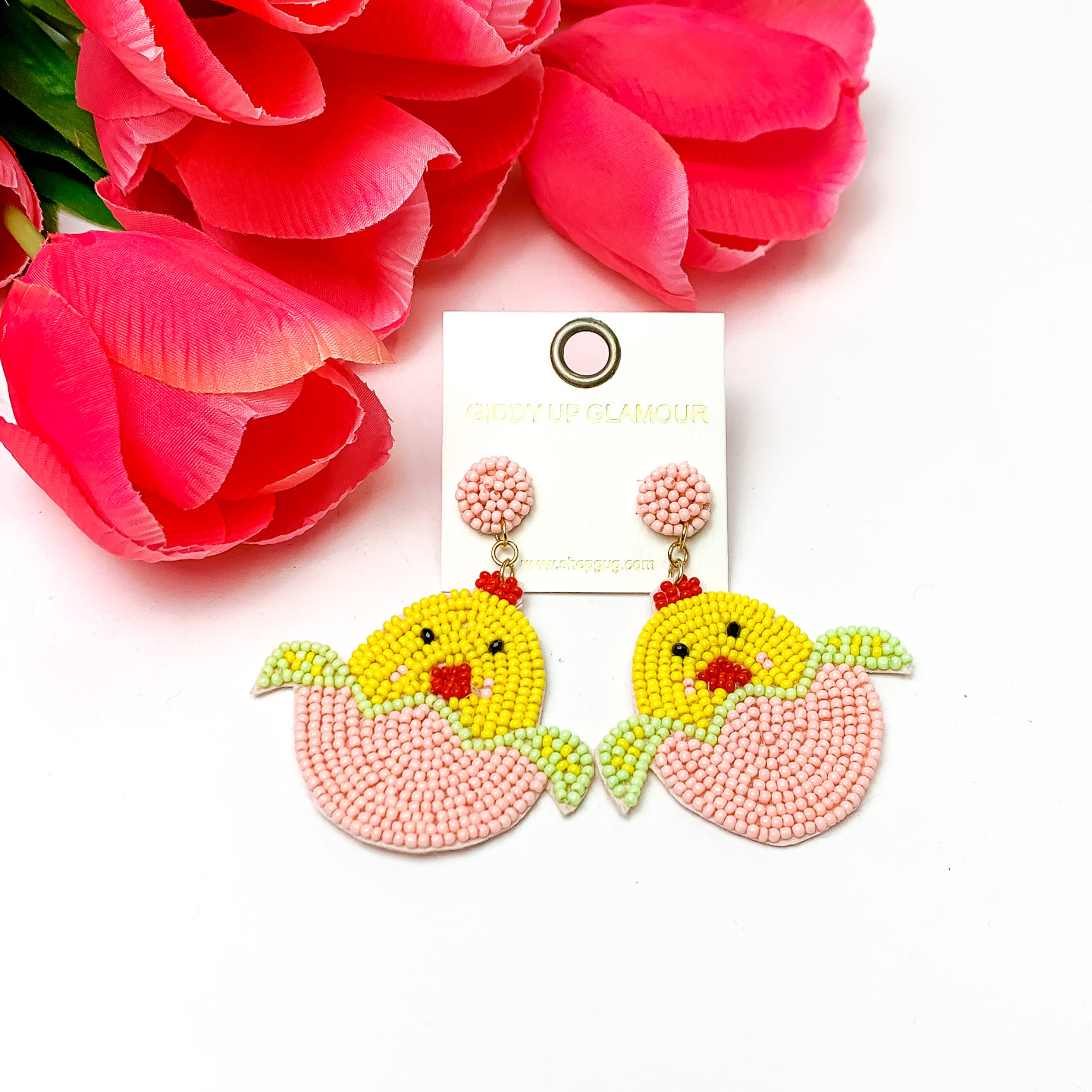 Beaded, light pink Easter egg earrings with a yellow beaded chick hatching. Pictured on white background with red-coral colored flowers.
