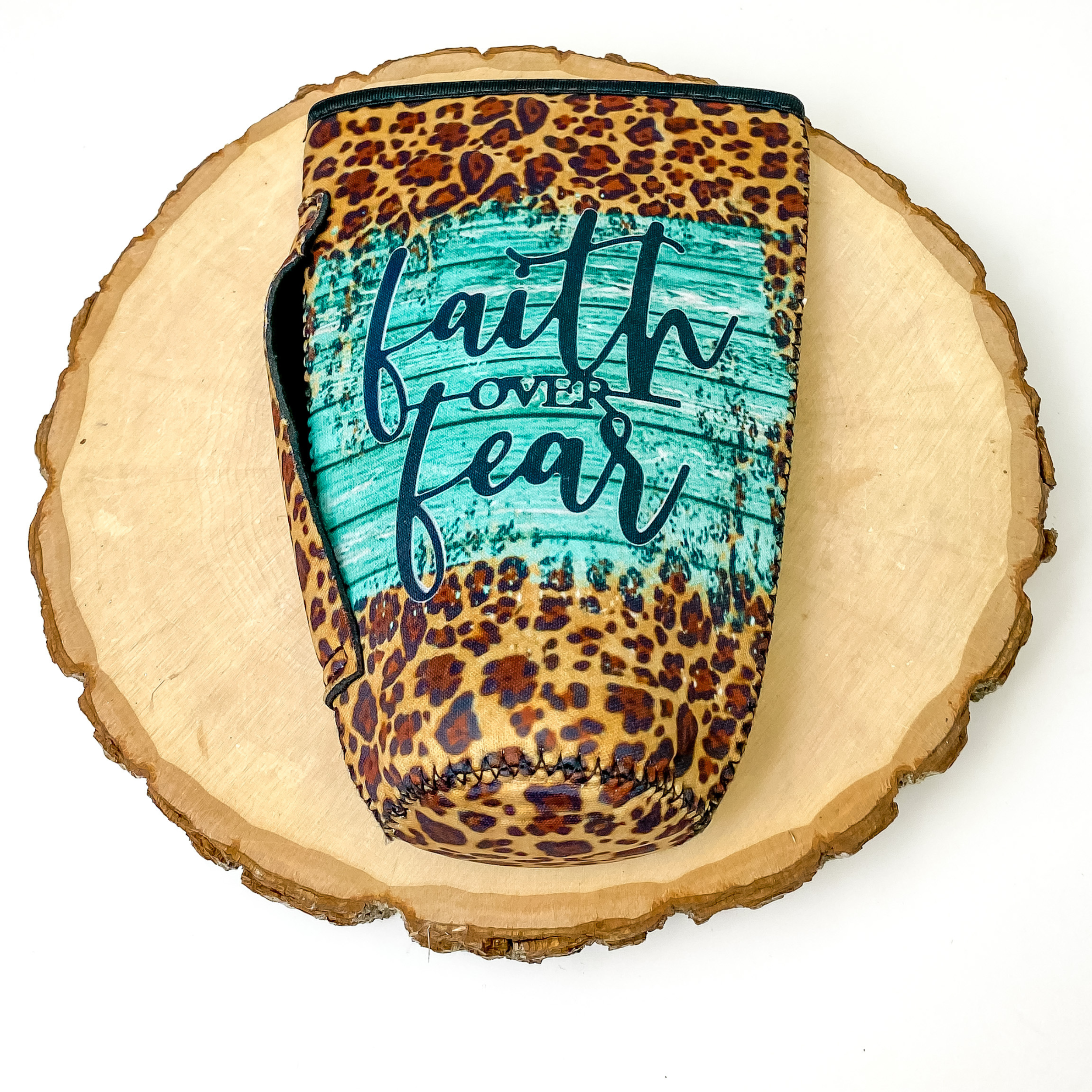 Tumbler drink sleeve with the saying "Faith over fear" with turquoise wood print behind it. This drink sleeve also includes a handle and leopard print all over. This drink sleeve is pictured on a piece of wood on a white background.