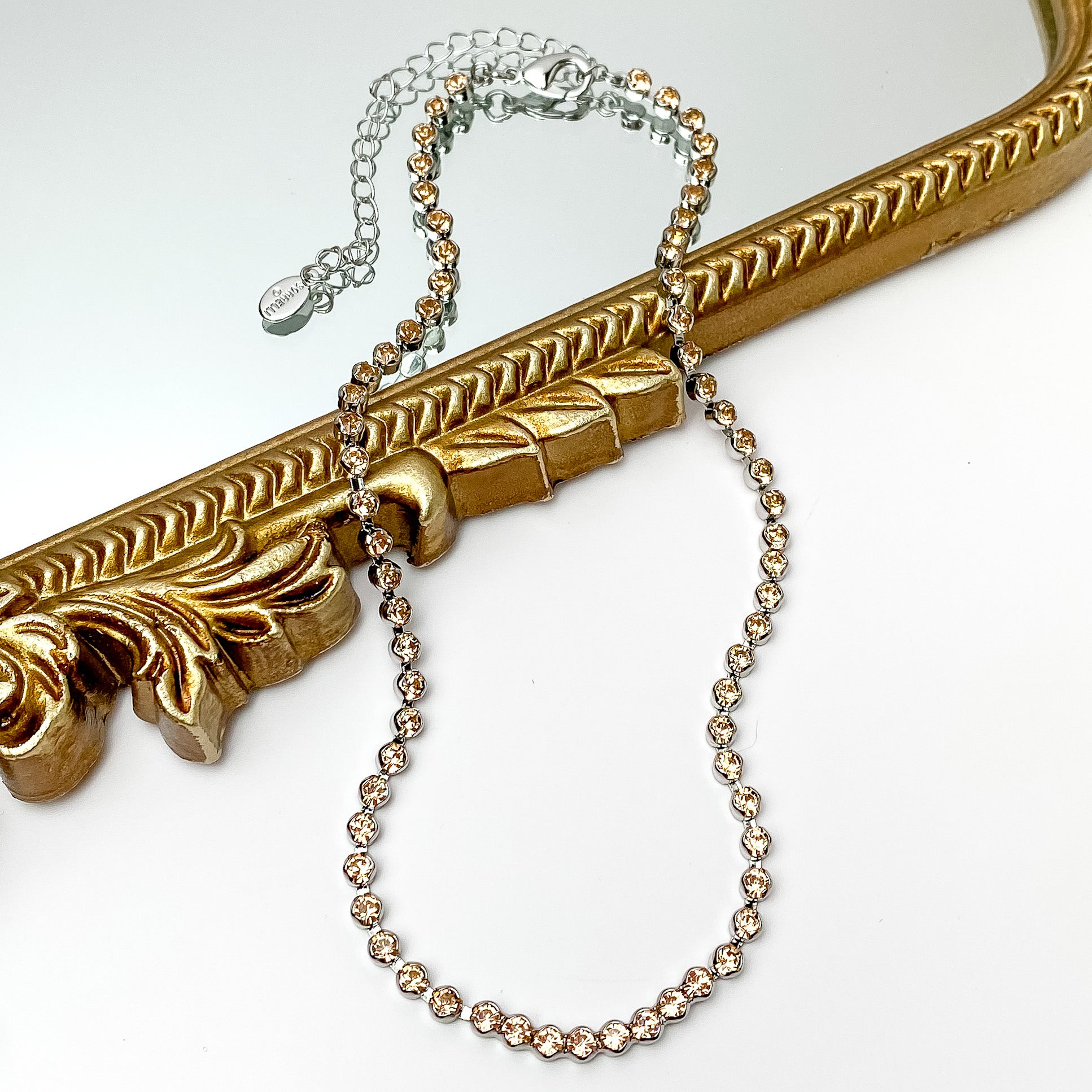Pictured is a silver necklace with light beige crystals. This necklace is pictured partially on a gold mirror on a white background.