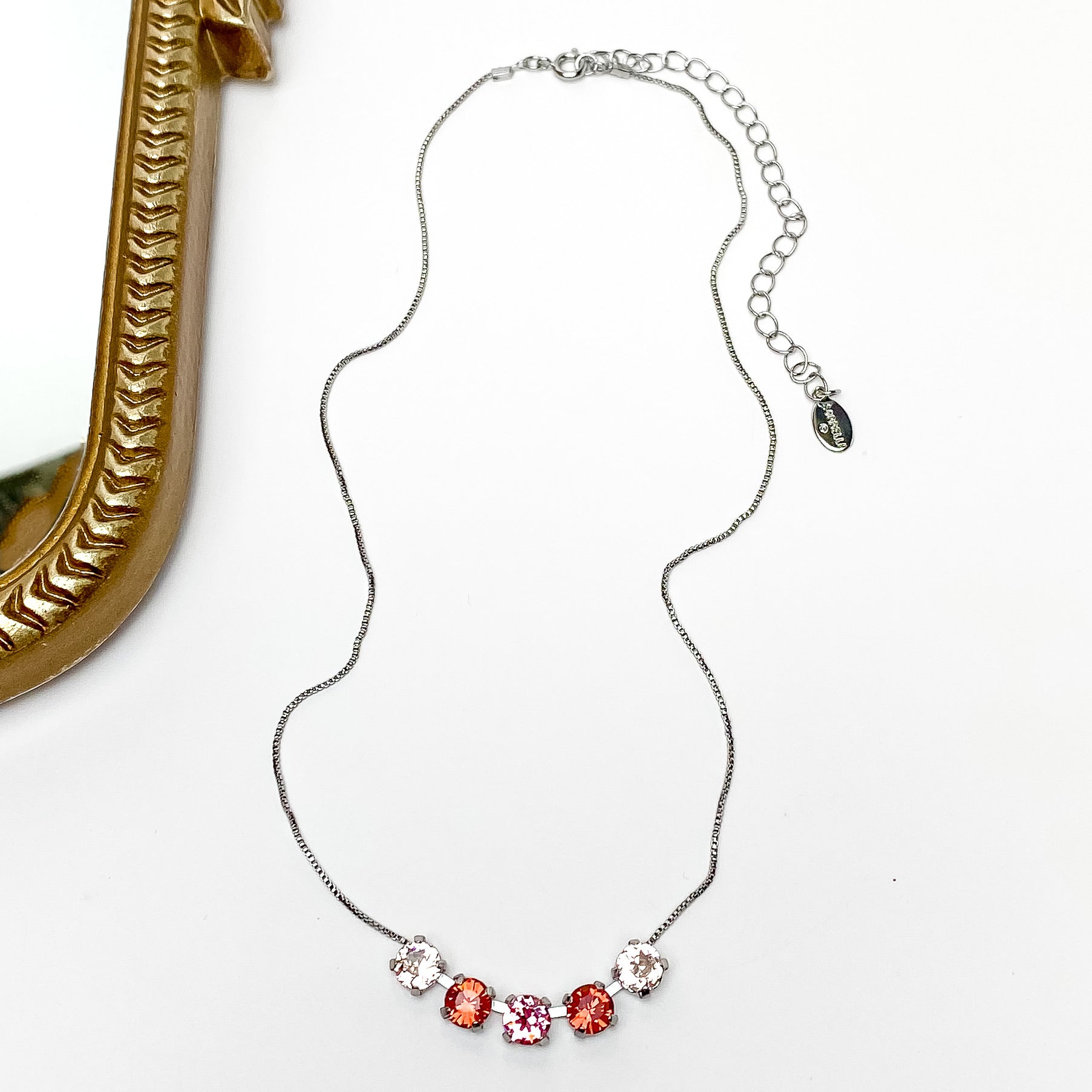 Pictured is a silver necklace with a five crystals. The center crystal is pink, the next one on each side is red, and finally the last two are a light champagne colored crystal. This necklace is pictured on a white background with a gold mirror on the left side.