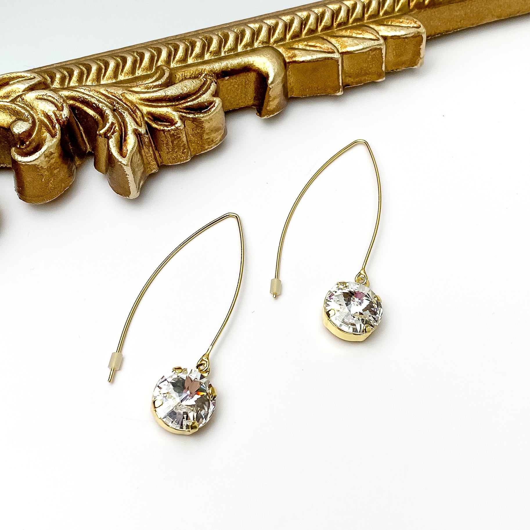Gold, kidney wire dangle earrings. These earrings include a round clear crystal drop. These earrings are pictured in front of a gold mirror on a white background. 
