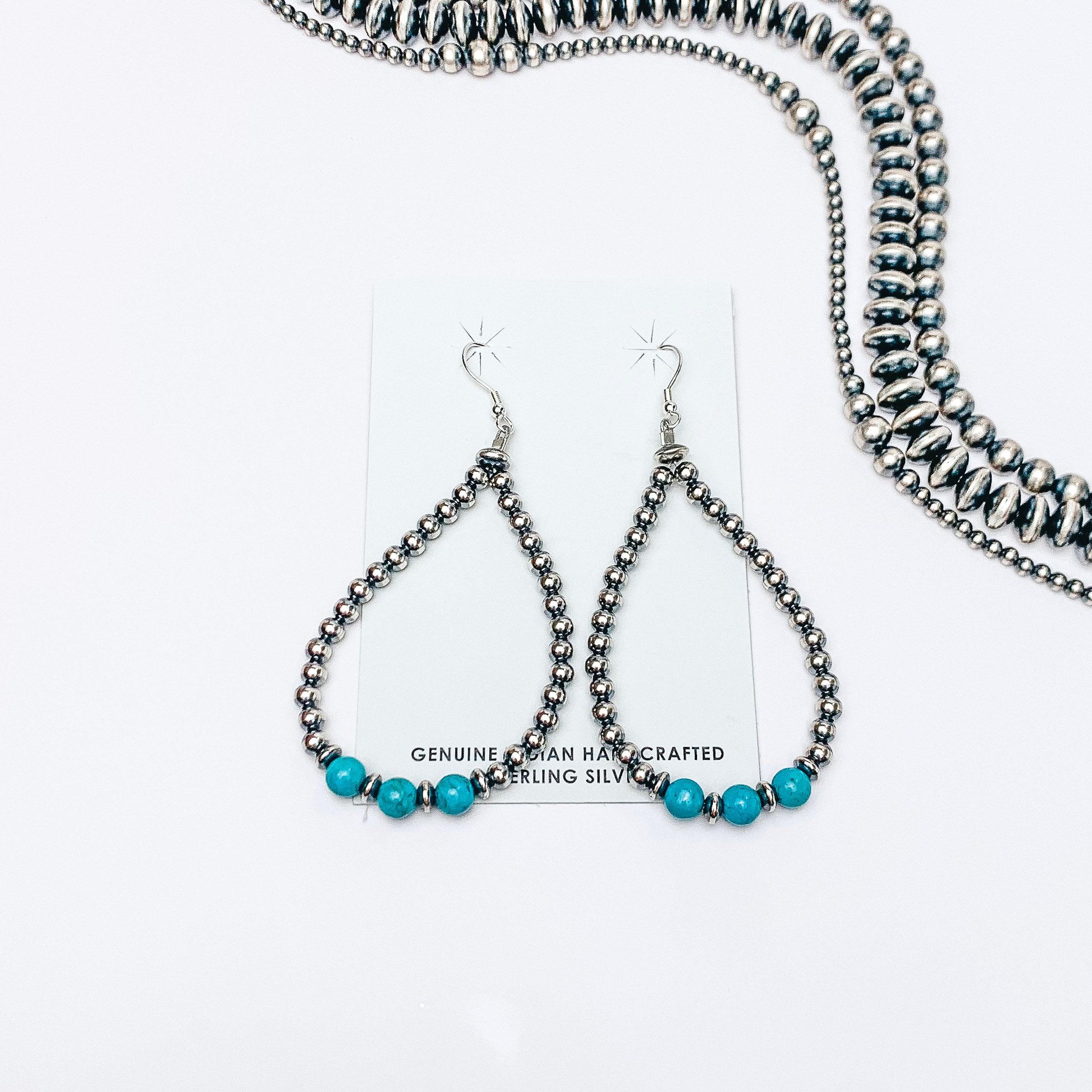 Centered in the picture is tear drop earrings made from  navajo pearls and sleeping beauty turquoise. Navajo pearls are laid above the earrings on white background. 