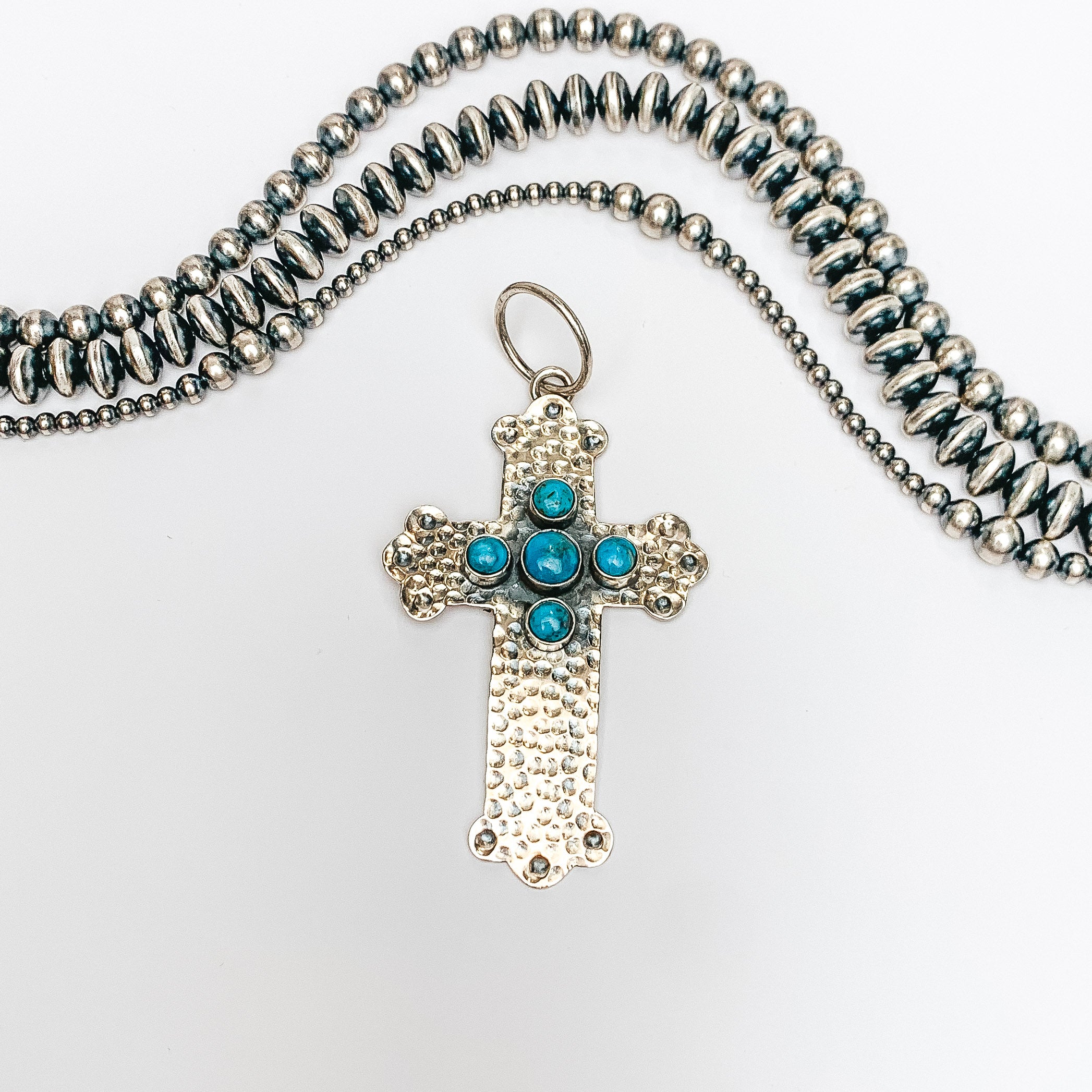 Centered in the picture is a hammered pattern cross pendant with 5 kingman turquoise stones. Navajo pearls are laid above the cross, all on a white background.