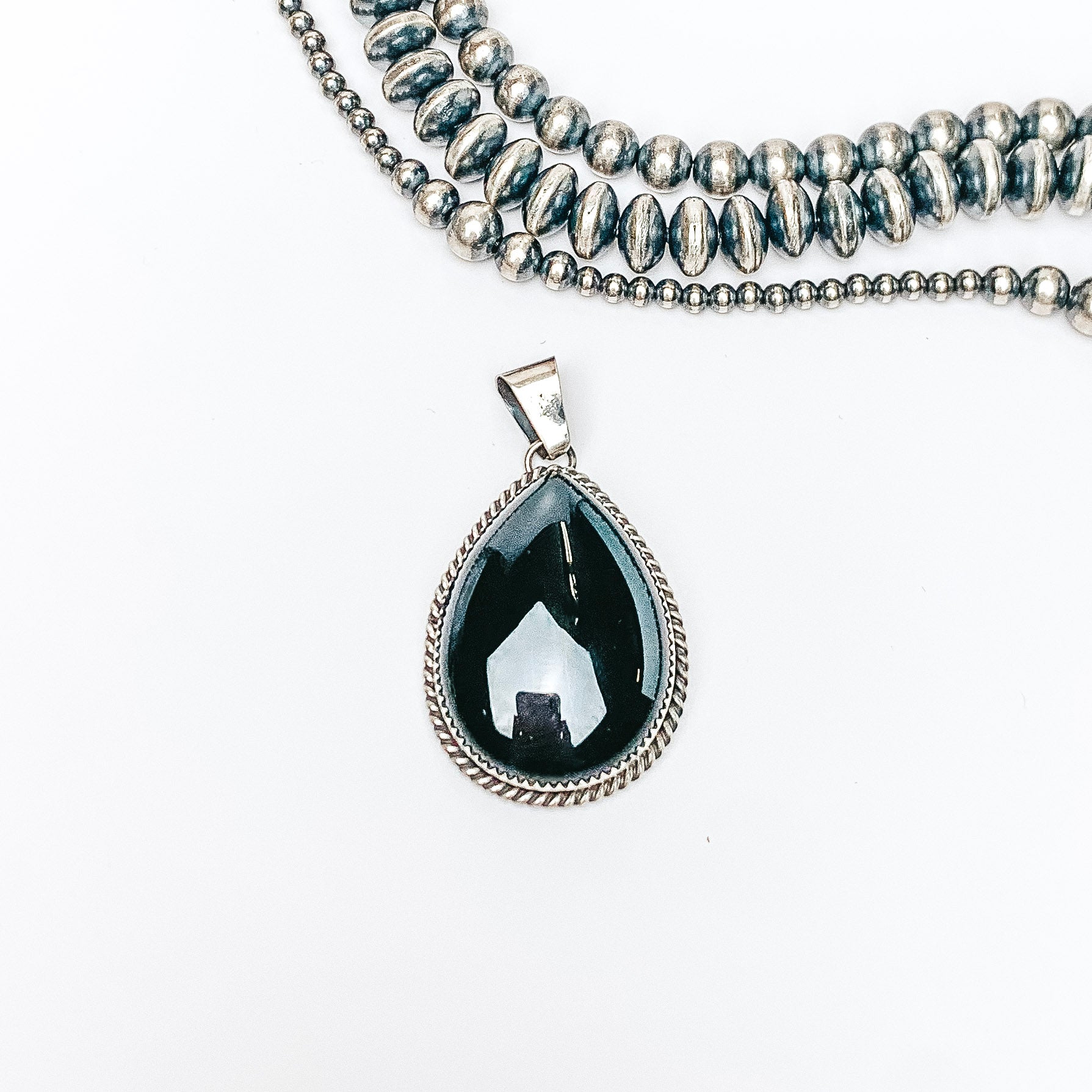 Centered in the picture is a black onyx teardrop pendant. Above the pendant is various sized navajo pearls. All on a white background. 