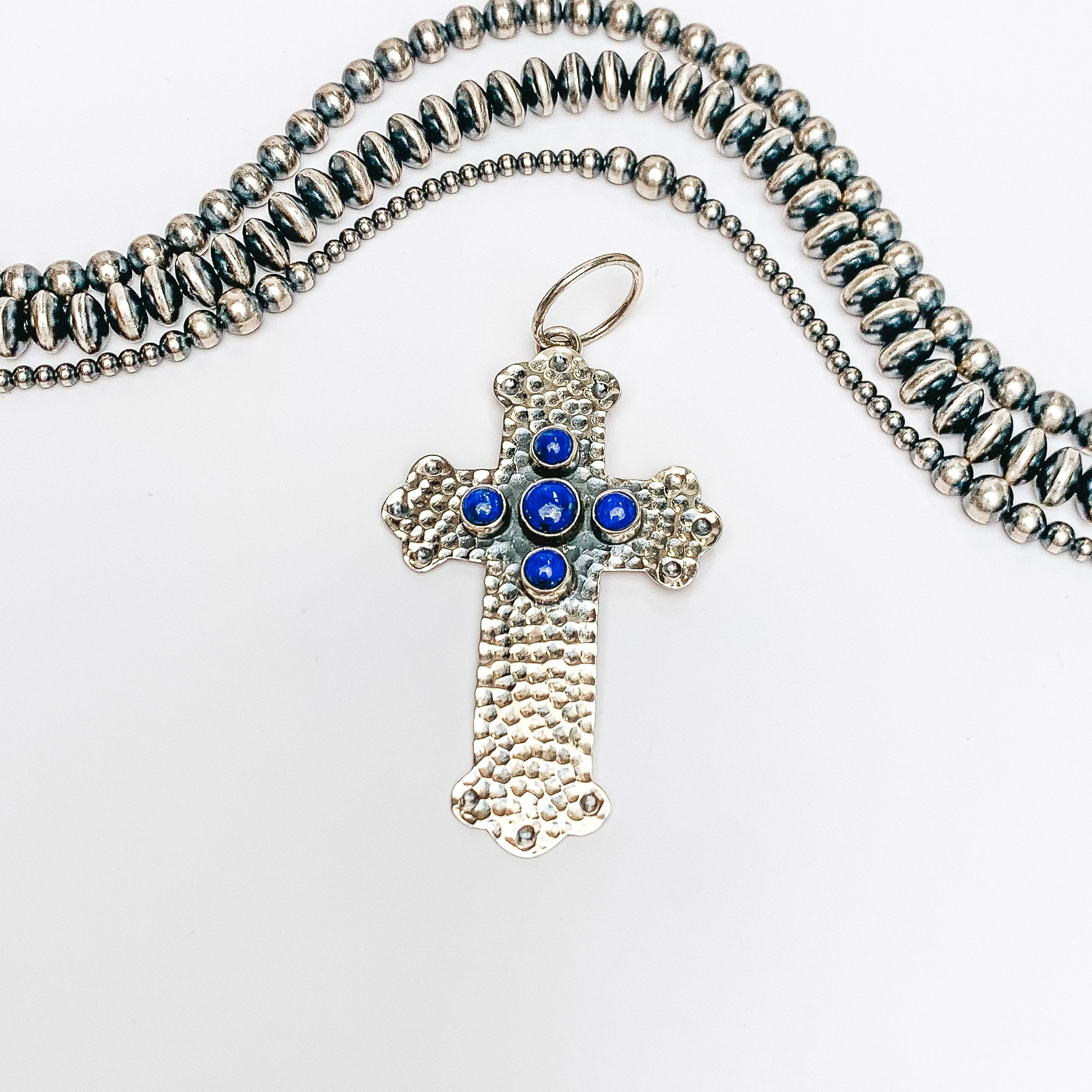 Centered in the picture is a hammered pattern cross pendant with 5 dark lapis stones. Navajo pearls are laid above the cross, all on a white background.