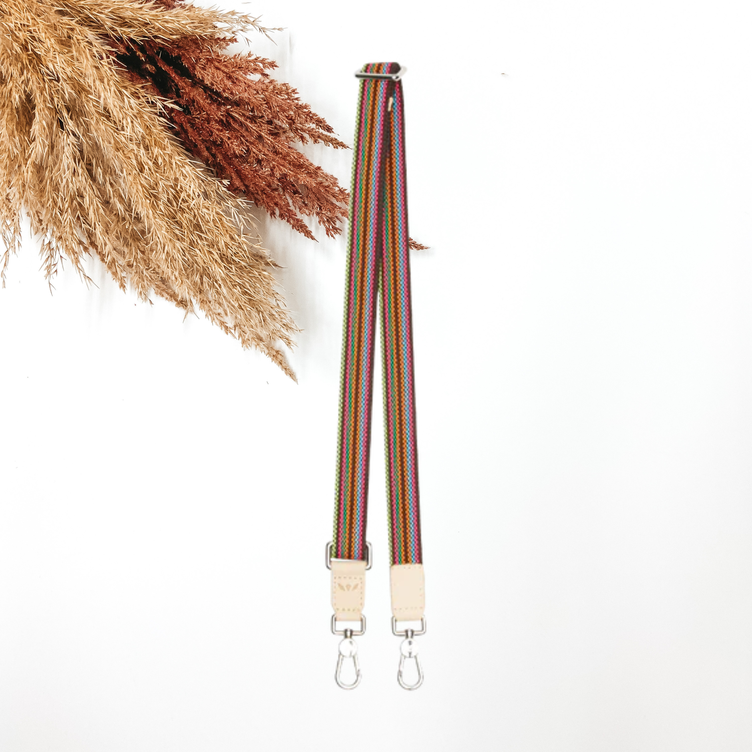 A woven multicolored purse strap that has a main color of brown. This purse strap is pictured on a white background with tan and brown pompous grass in the top left corner.
