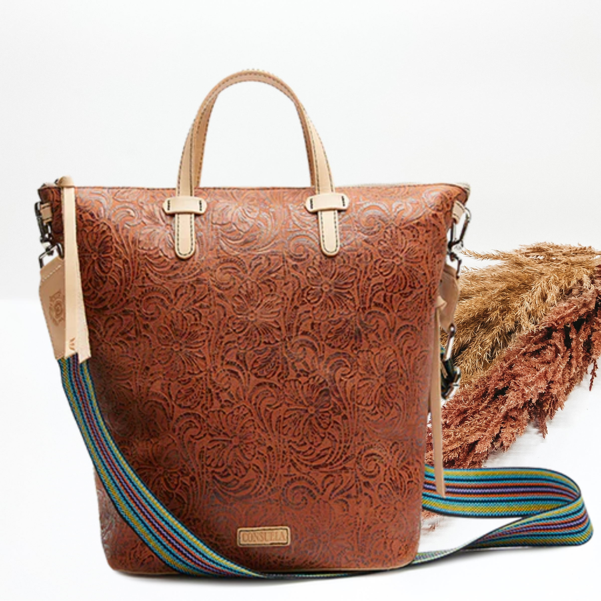 Pictured is a sling bag with a brown tooled print. This bag inlcudes a top zipper with short light tan straps and a thick striped strap. This bag is pictured on a white background with pompous grass on the left side of the picture. 