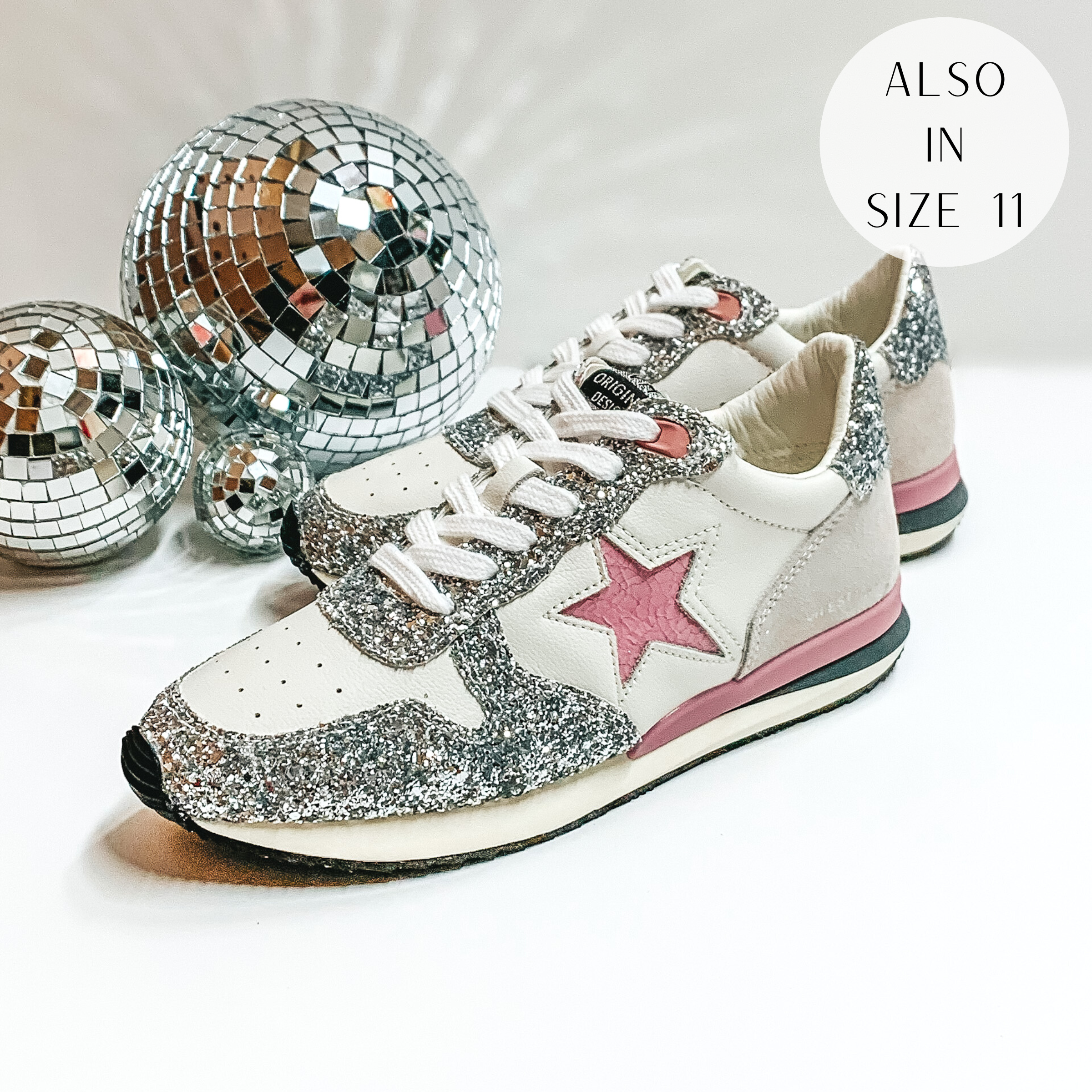 White tennis shoes with silver glitter design over the shoes. These shoes also have a pink star emblem, and a pink and grey part of the sole. These shoes are pictured on a white background with disco balls on the left hand side.