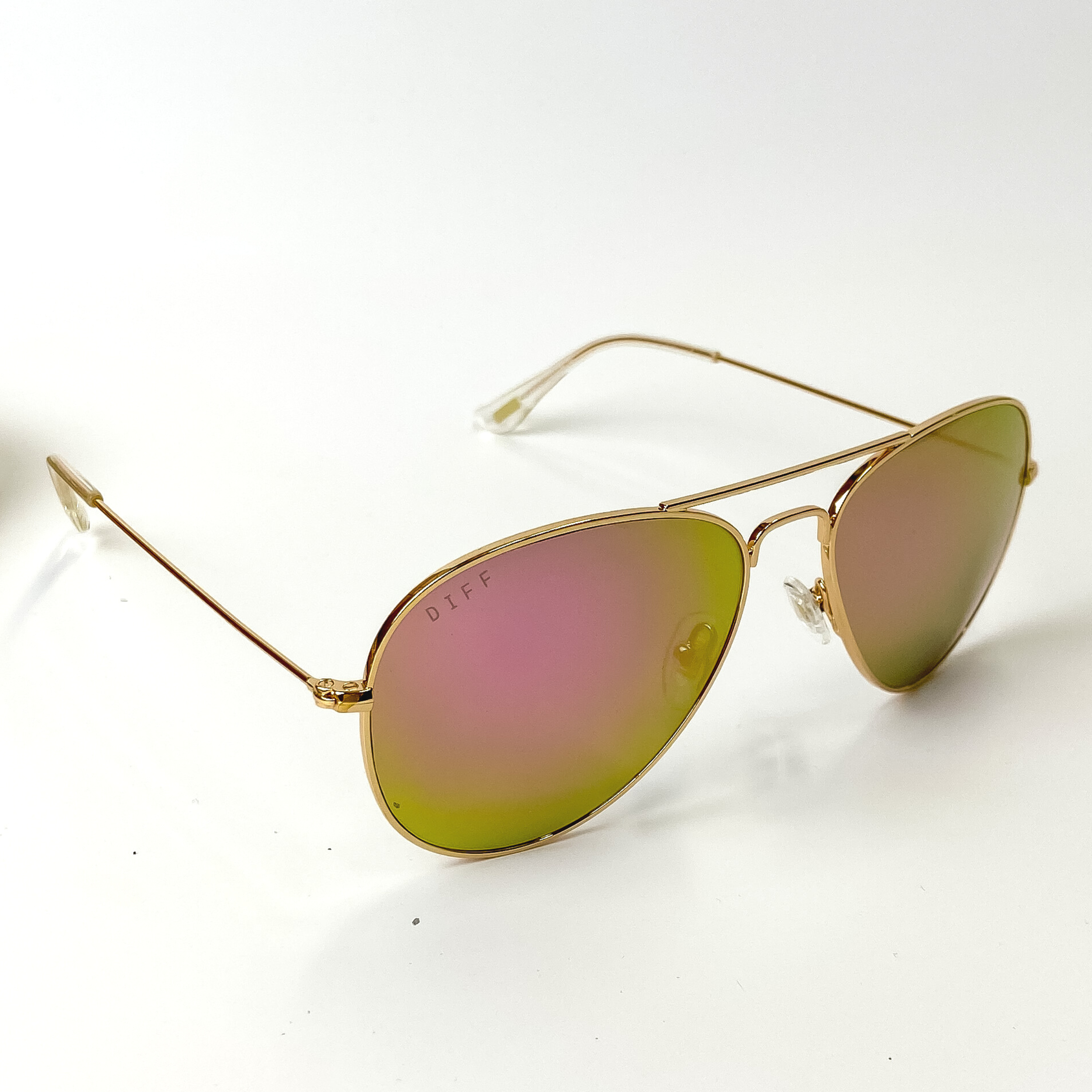 A pair of aviator style sunglasses with pink mirror lenses and gold tone frames. These sunglasses are pictured on a white background with gold jewelry.